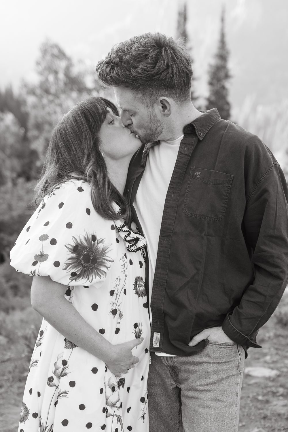 Black and white photo of a pregnant couple kissing outdoors, woman in polka dot dress cradling baby bump.
