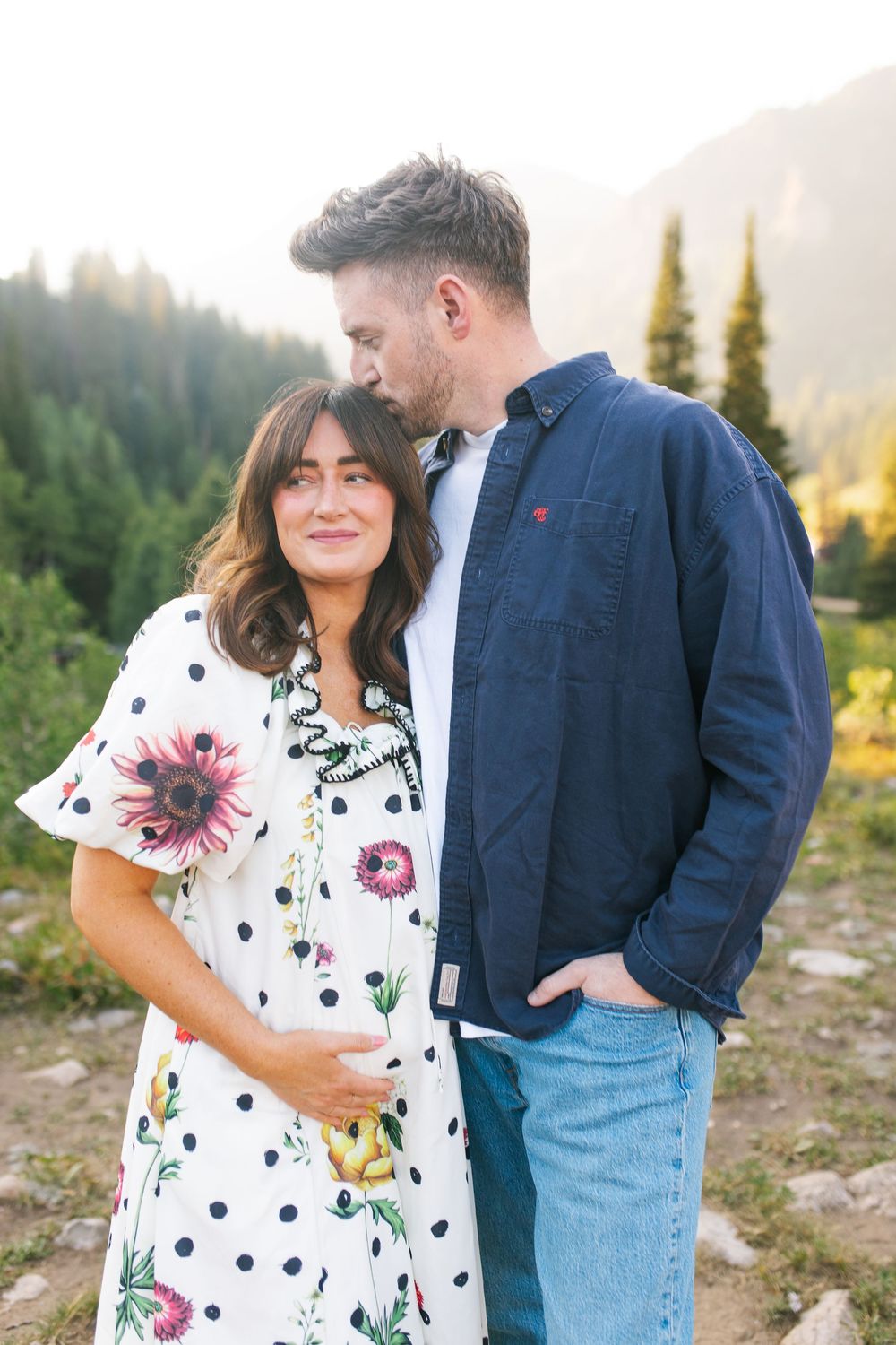Couple embracing outdoors in mountains, woman in floral polka dot dress, man in navy shirt kissing her head.