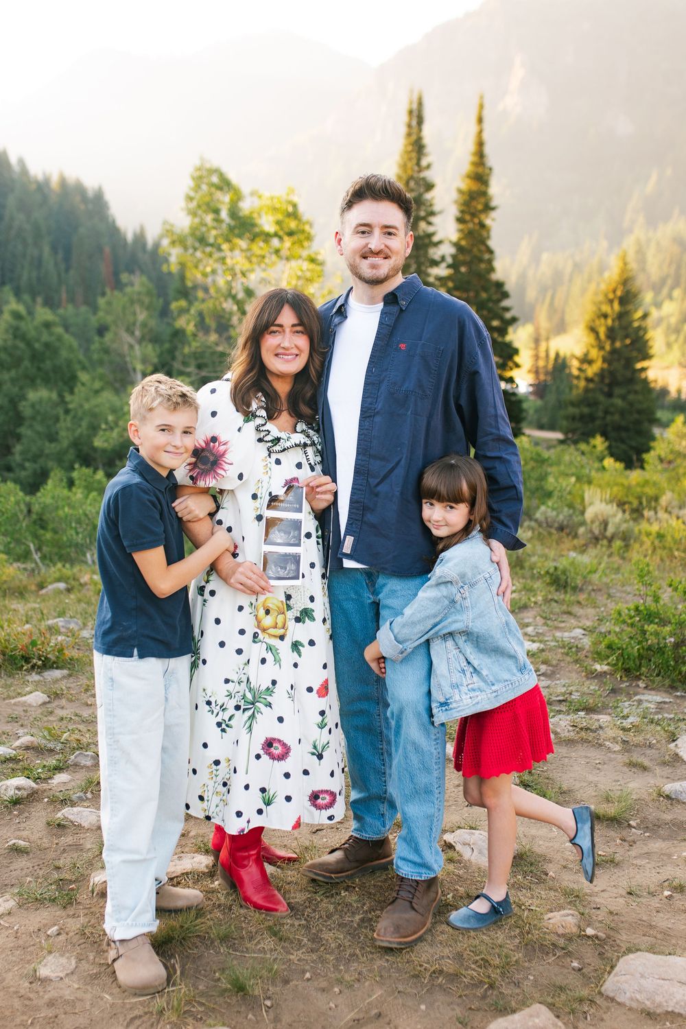 Happy family of four poses outdoors in mountain setting, pregnant mother holding ultrasound photos.