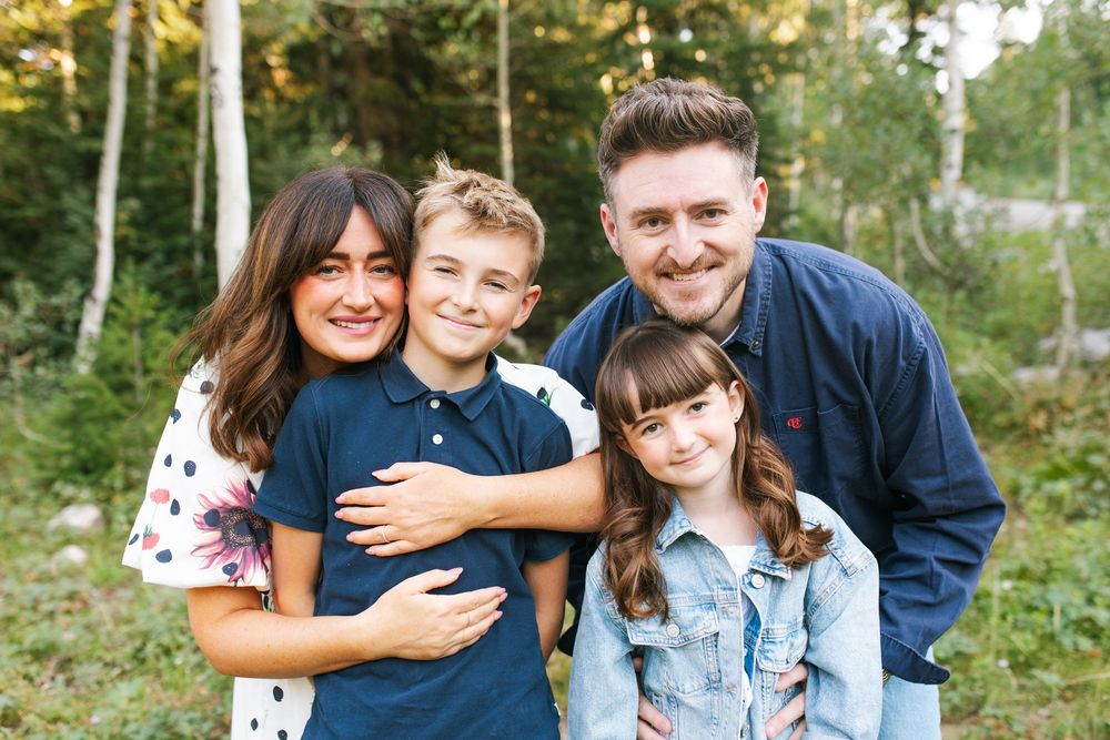 Happy family of four smiling outdoors in a forest setting, parents with two children posing for a portrait.