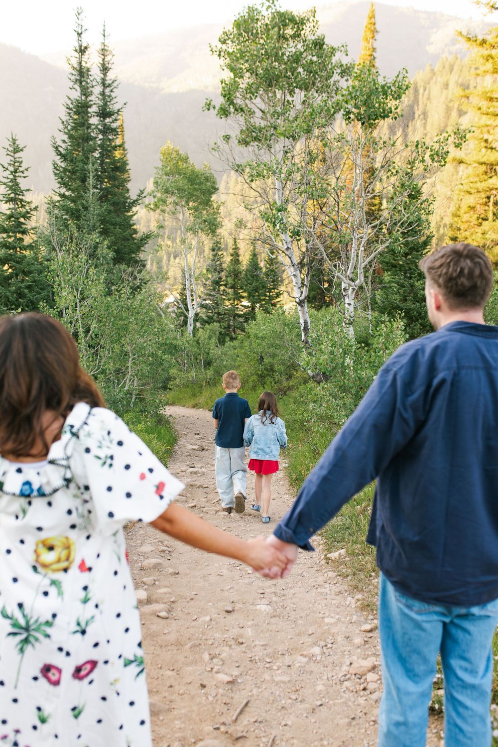 Family holding hands on a scenic mountain trail surrounded by lush green trees and autumn foliage.