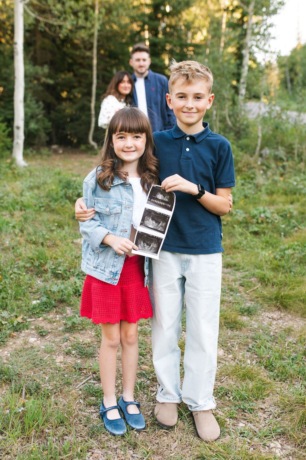 Two children proudly hold an ultrasound photo outdoors, announcing a pregnancy with parents standing behind them.
