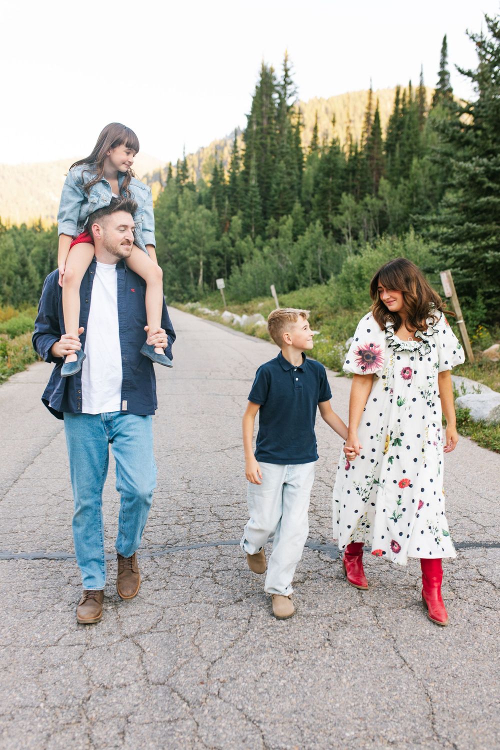 Family of four walking on mountain road, dad carrying girl on shoulders, mom in floral dress and red boots.