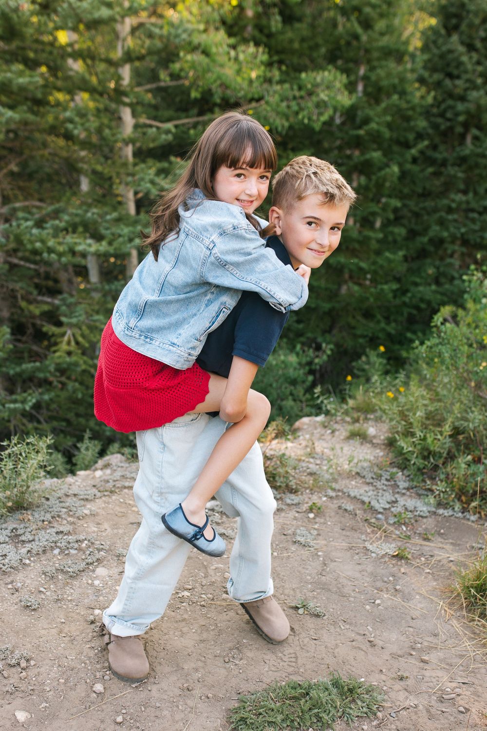 Boy giving younger girl a piggyback ride outdoors on a rocky trail surrounded by green trees.