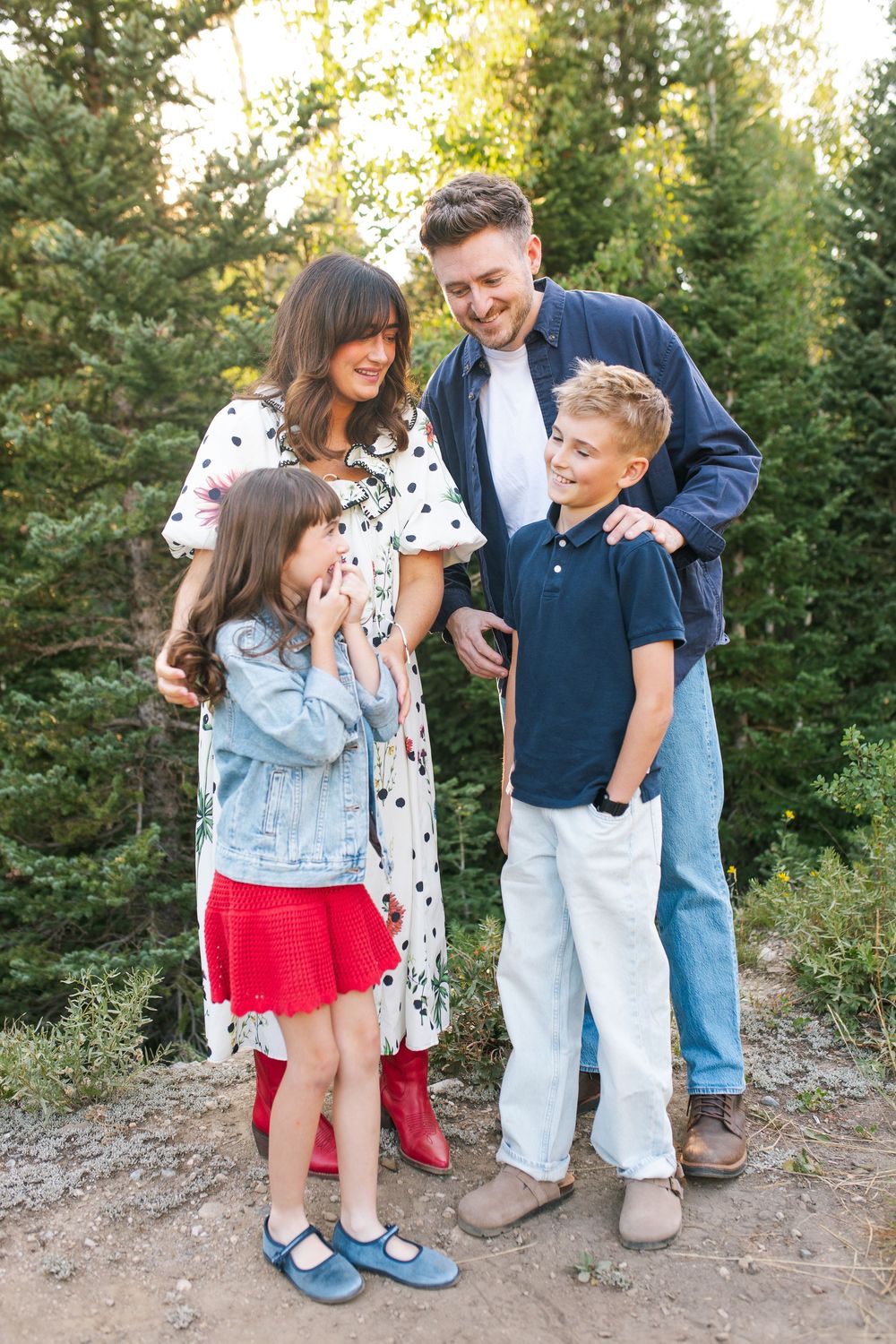 Happy family of four posing outdoors among pine trees, parents smiling at children in casual summer attire.