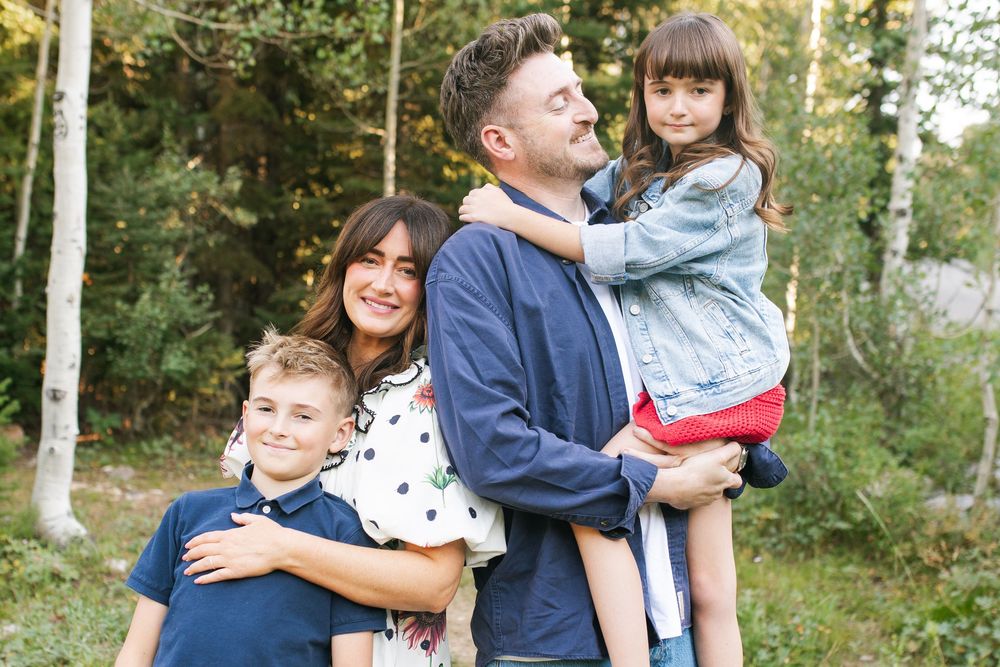 Happy family of four posing outdoors among trees, parents smiling with two young children in a warm, natural setting.