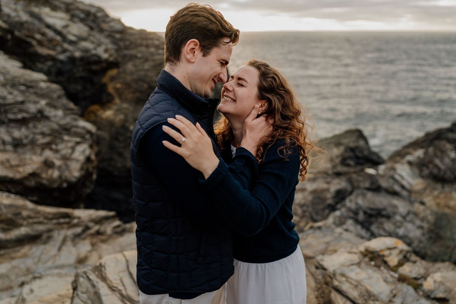 Couple embracing romantically on rocky coastal cliffs with ocean waves in the background during golden hour.