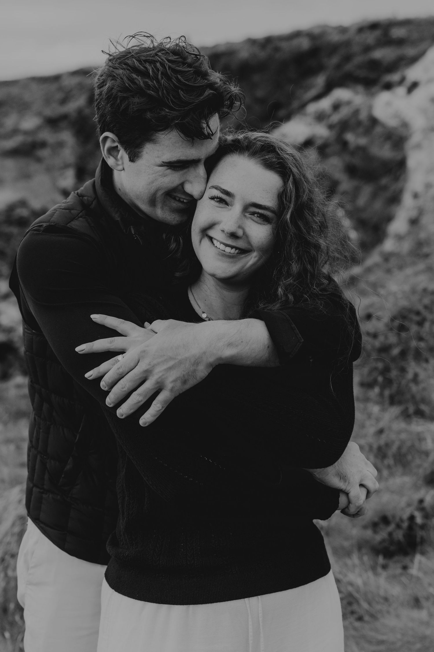Black and white photo of a smiling couple embracing outdoors, with rocky landscape in the background at trevose head