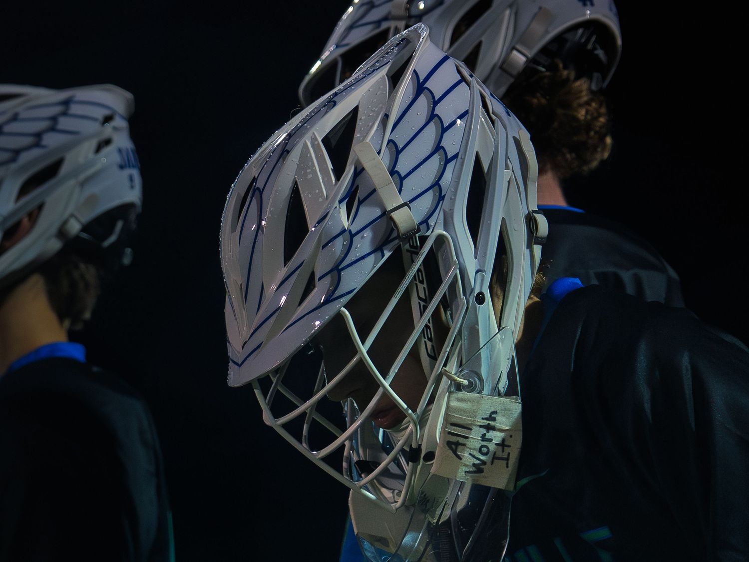 A lacrosse player in a white winged helmet beaded with water bows their head in low blue light, helmeted teammates flanking them at a game