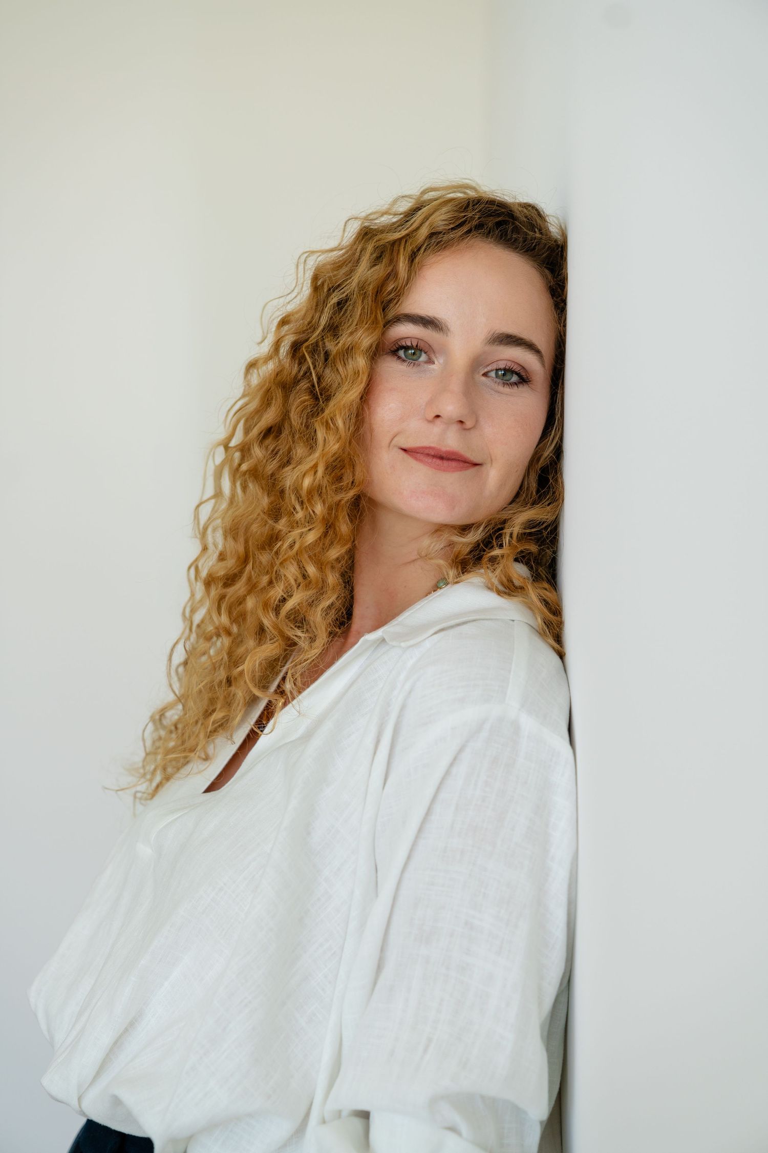 Young woman with curly blonde hair and blue eyes leaning against a white wall in a casual white linen top.
