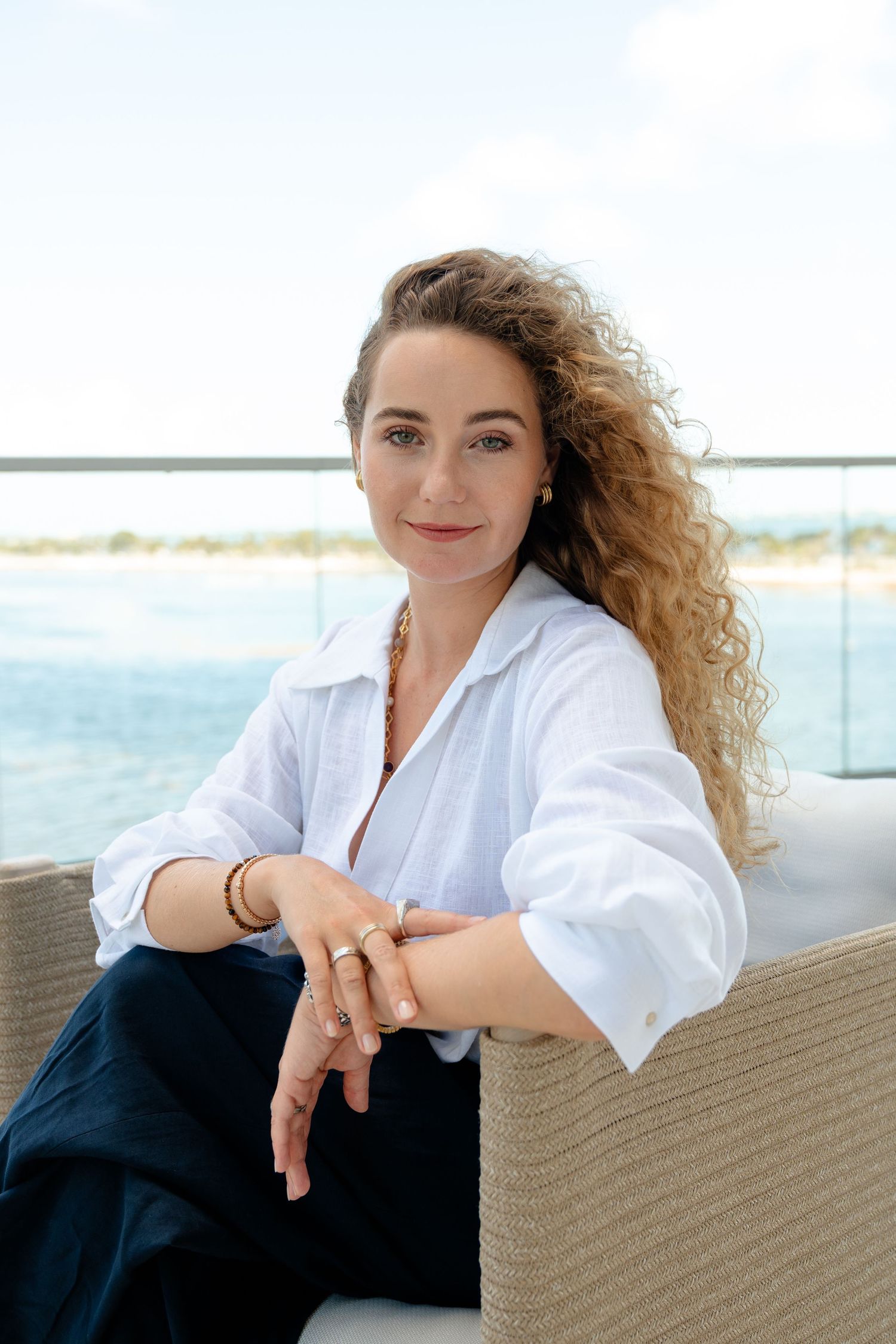 Woman with curly hair in white shirt sits relaxed on wicker chair with ocean view backdrop.