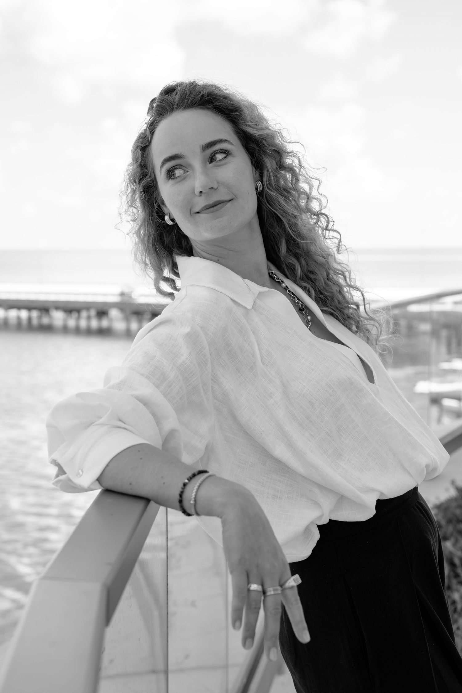 Black and white portrait of a curly-haired woman in white linen shirt leaning on railing by the sea.