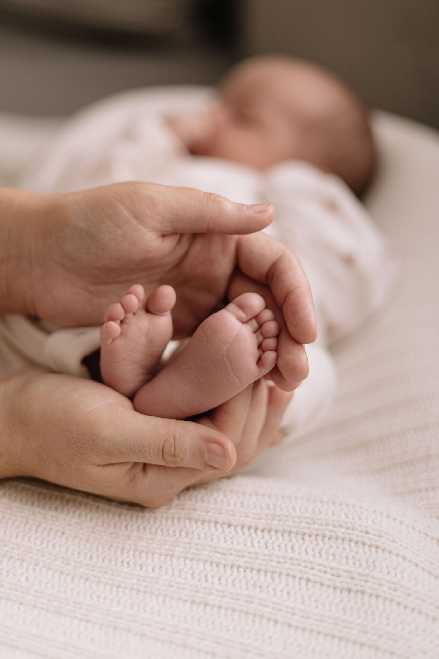 Adult hands gently cradle the tiny bare feet of a sleeping newborn baby wrapped in soft white swaddle on a cozy knit blanket.