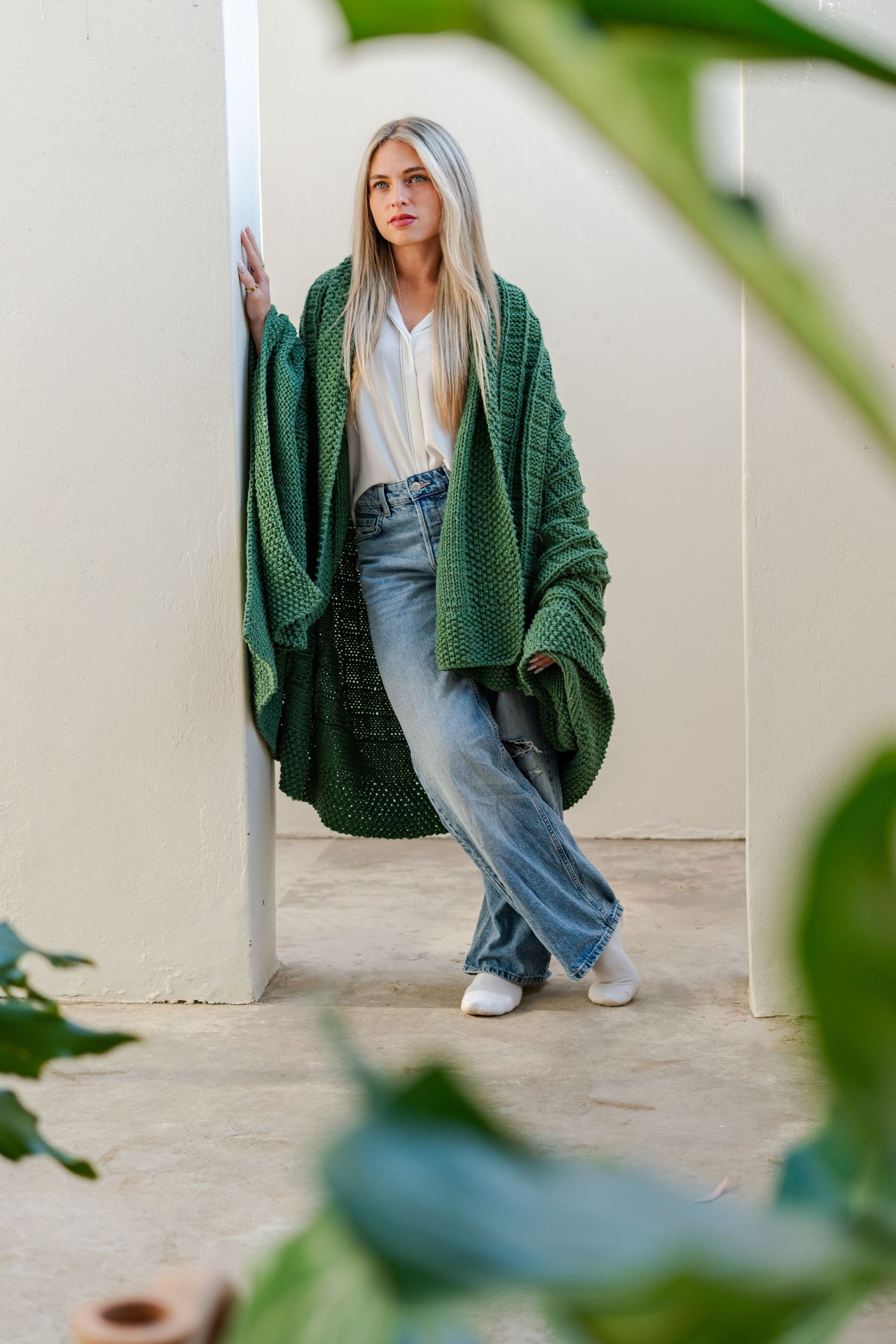 Woman in green knit cardigan, white blouse, and wide-leg jeans leaning against a white pillar outdoors during a brand photoshoot.
