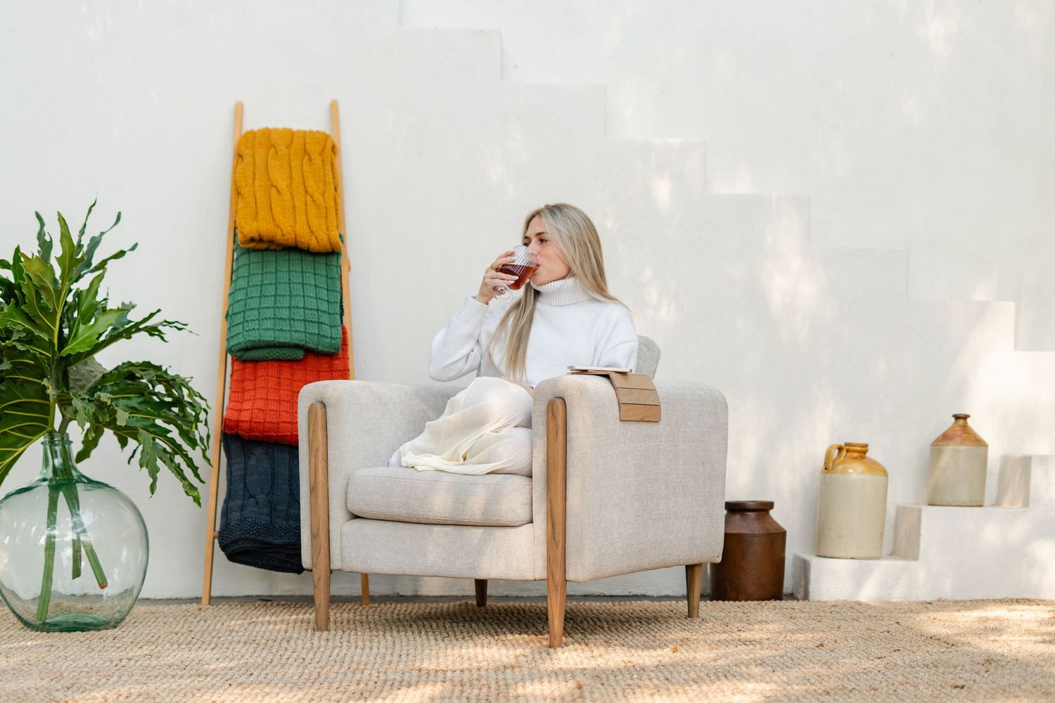 Lifestyle product photography of a woman relaxing in a bespoke beige armchair, sipping a drink.