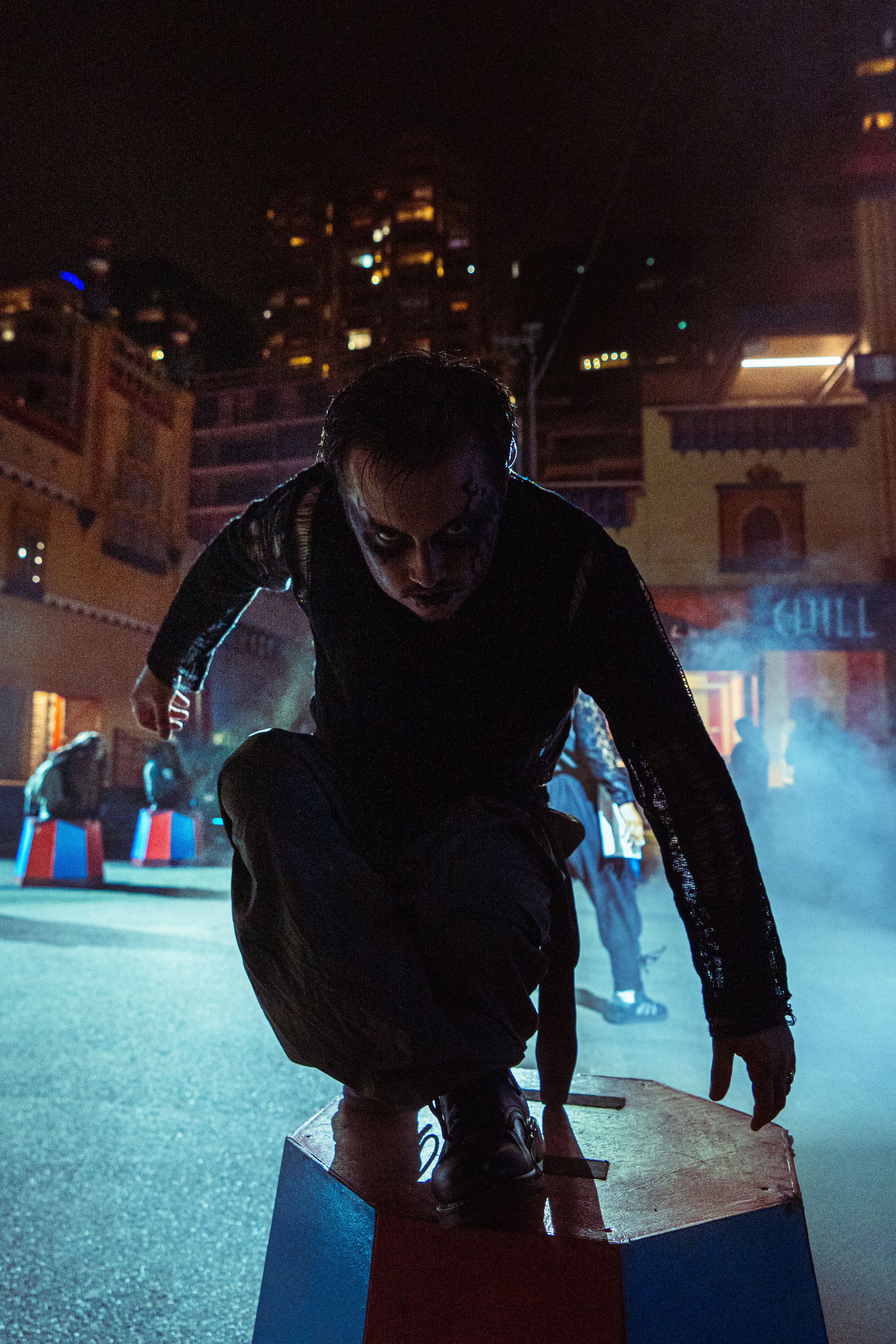 Person in dark costume with dramatic face paint crouches on colorful block at night event.