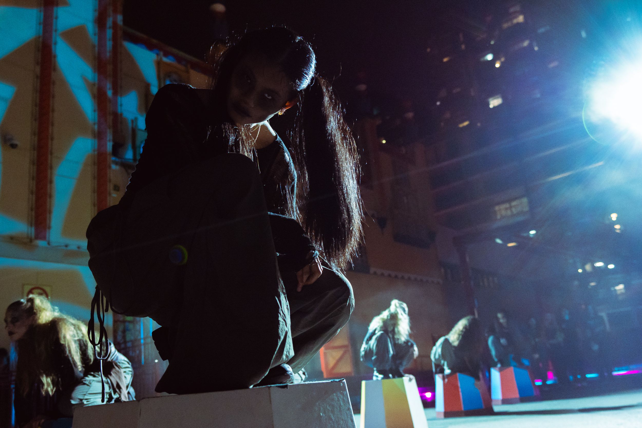 Person leaning over illuminated display in dark dramatic stage setting with colorful lighting.