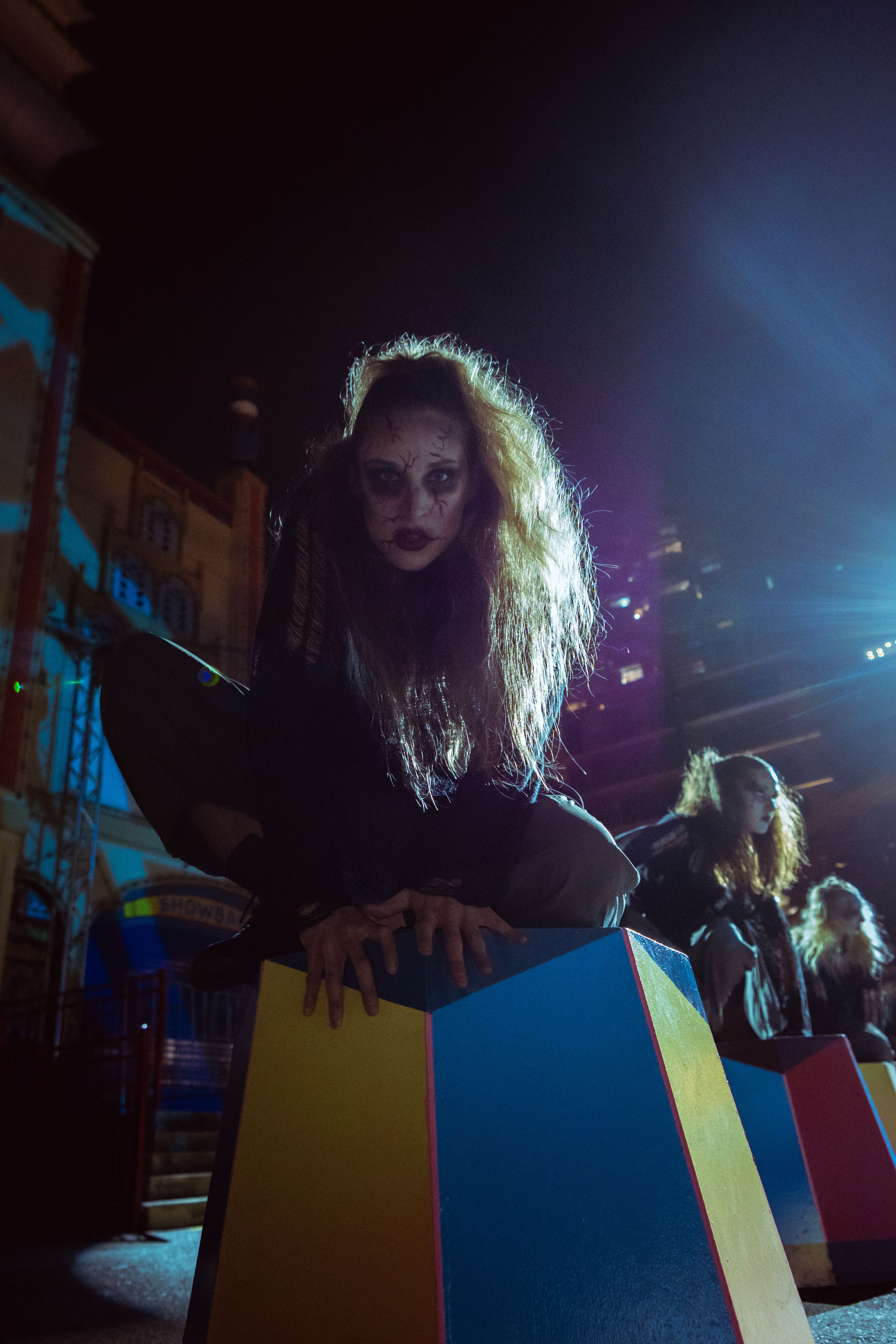 Woman with wild hair and dark makeup leans over colorful blocks in dramatic night setting.