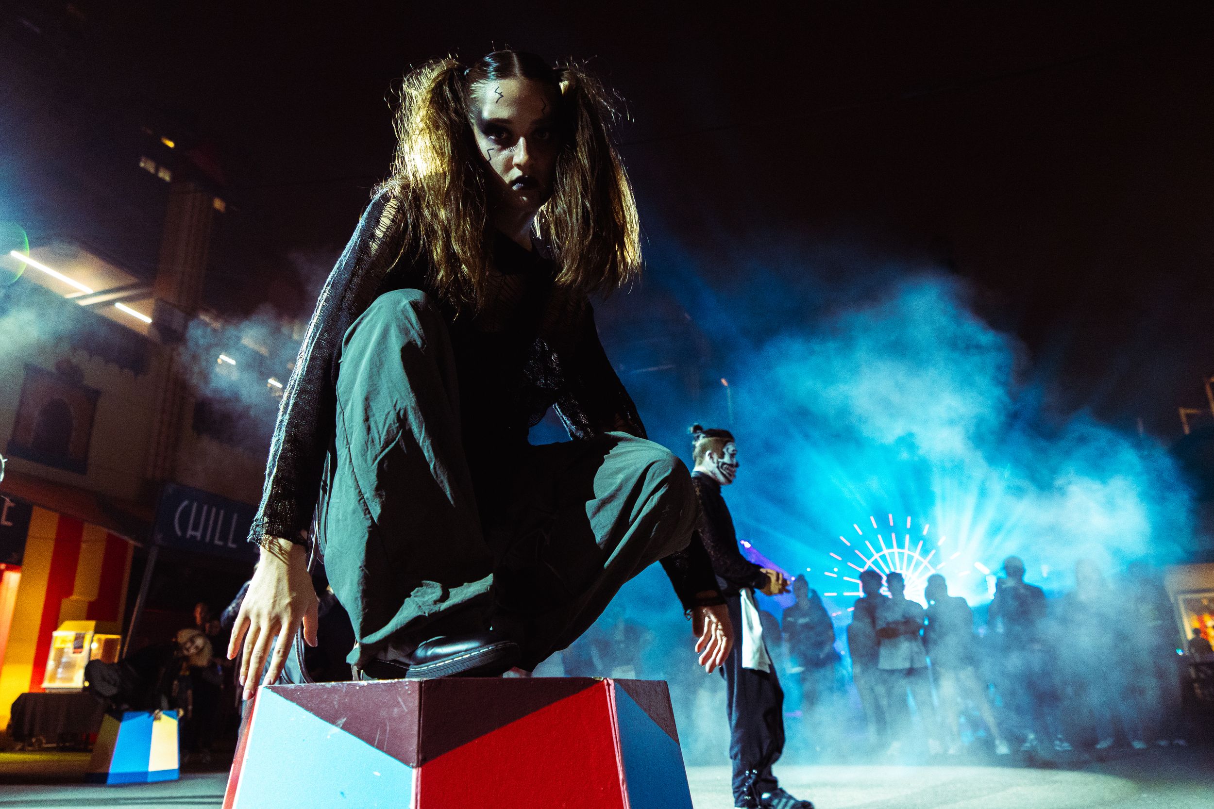 Long-haired performer in black outfit poses dynamically on colorful block with blue smoke backdrop.