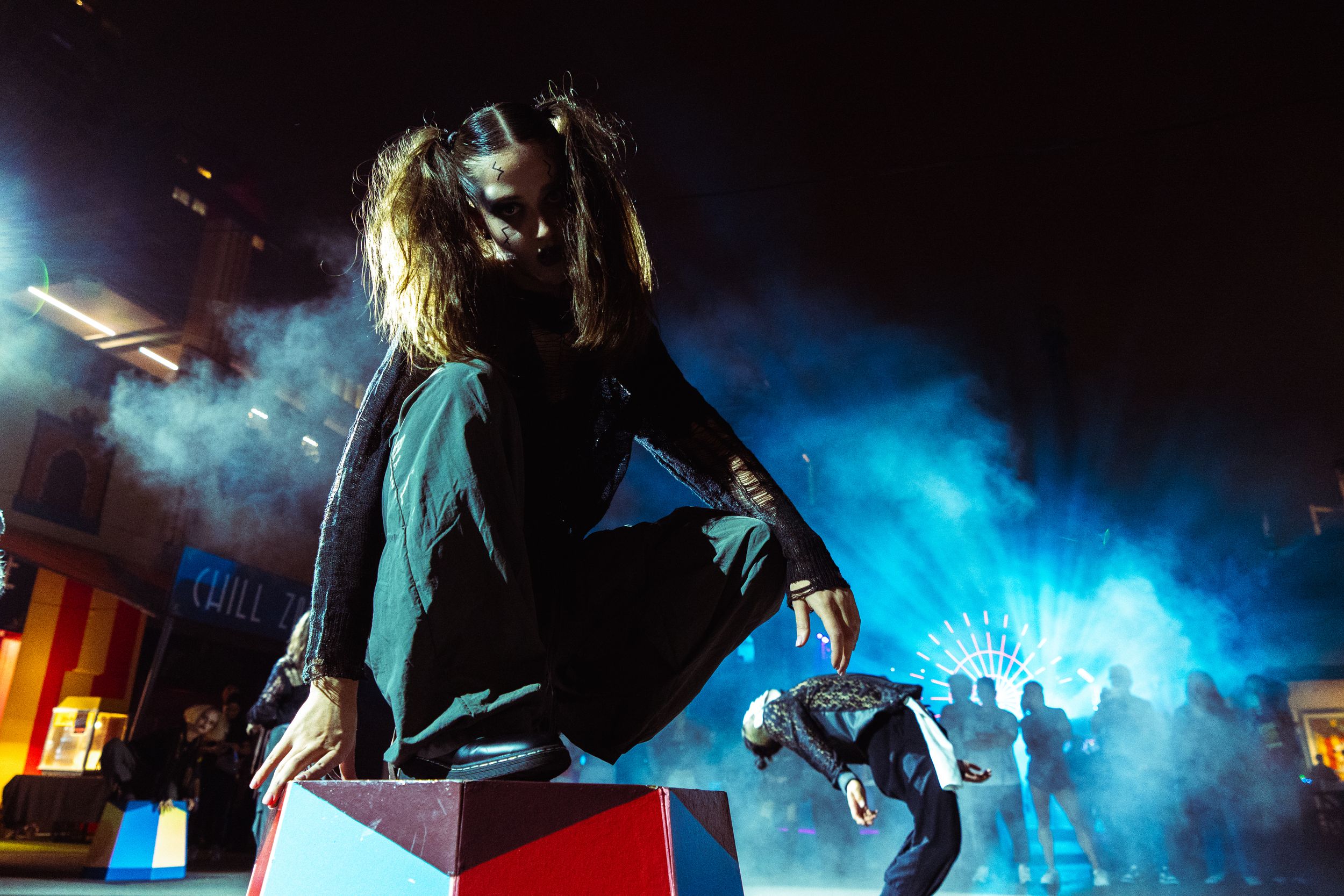 Performer with long hair leaps over colorful block against dramatic blue smoky night backdrop.
