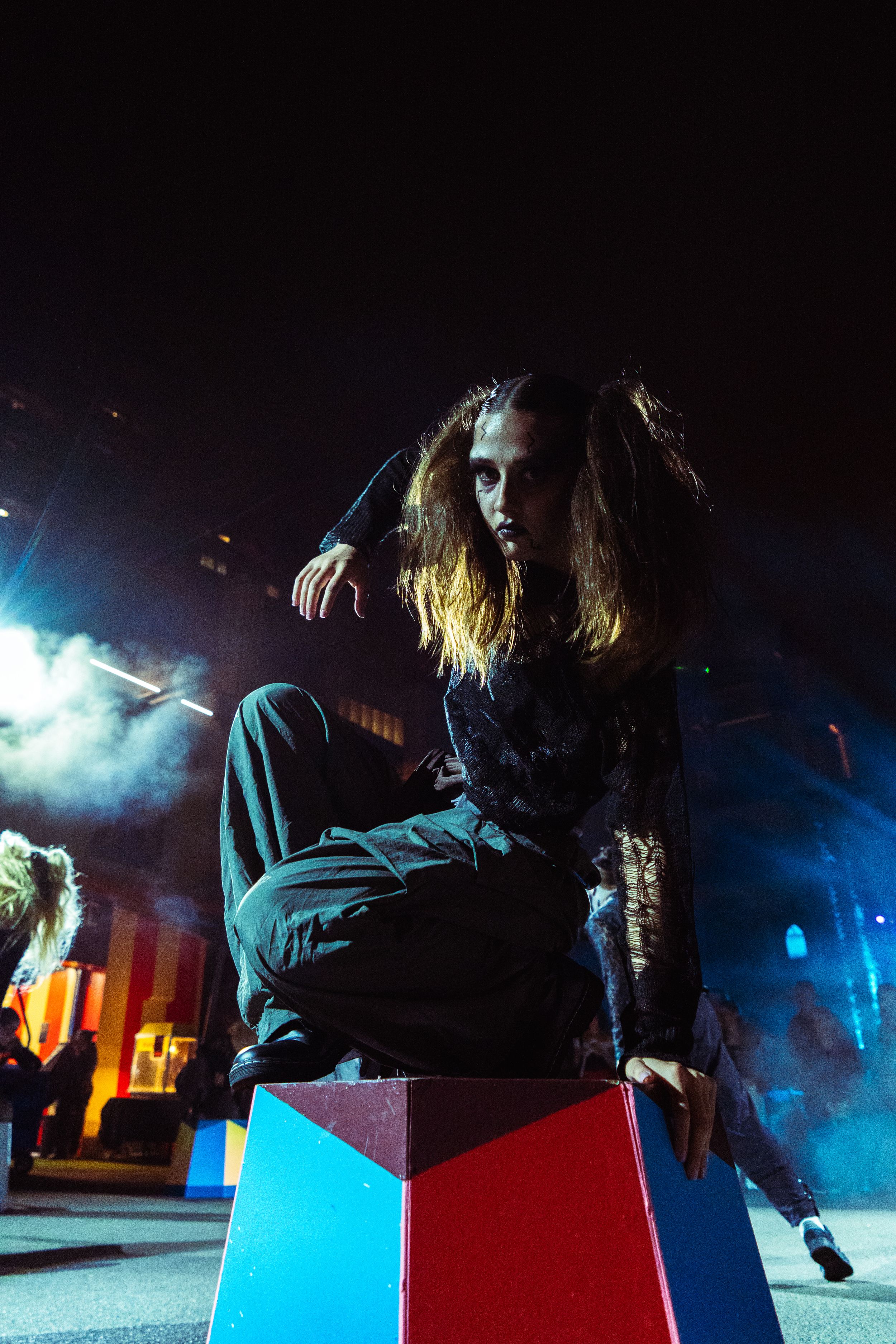 Long-haired person in black outfit sits on colorful block under dramatic night event lighting.