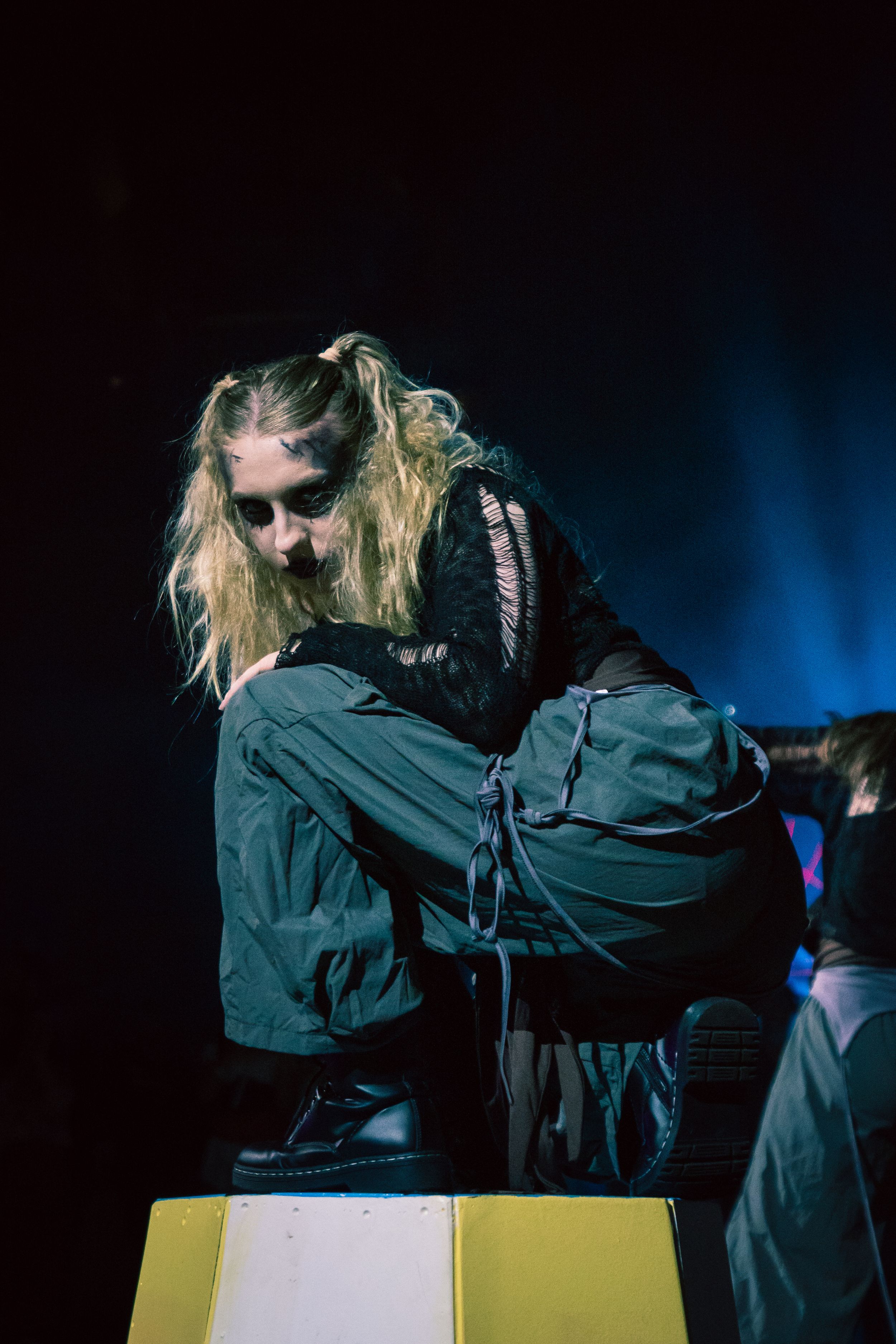 A performer with long blonde hair crouches on a yellow box under dramatic stage lighting in dark clothing.