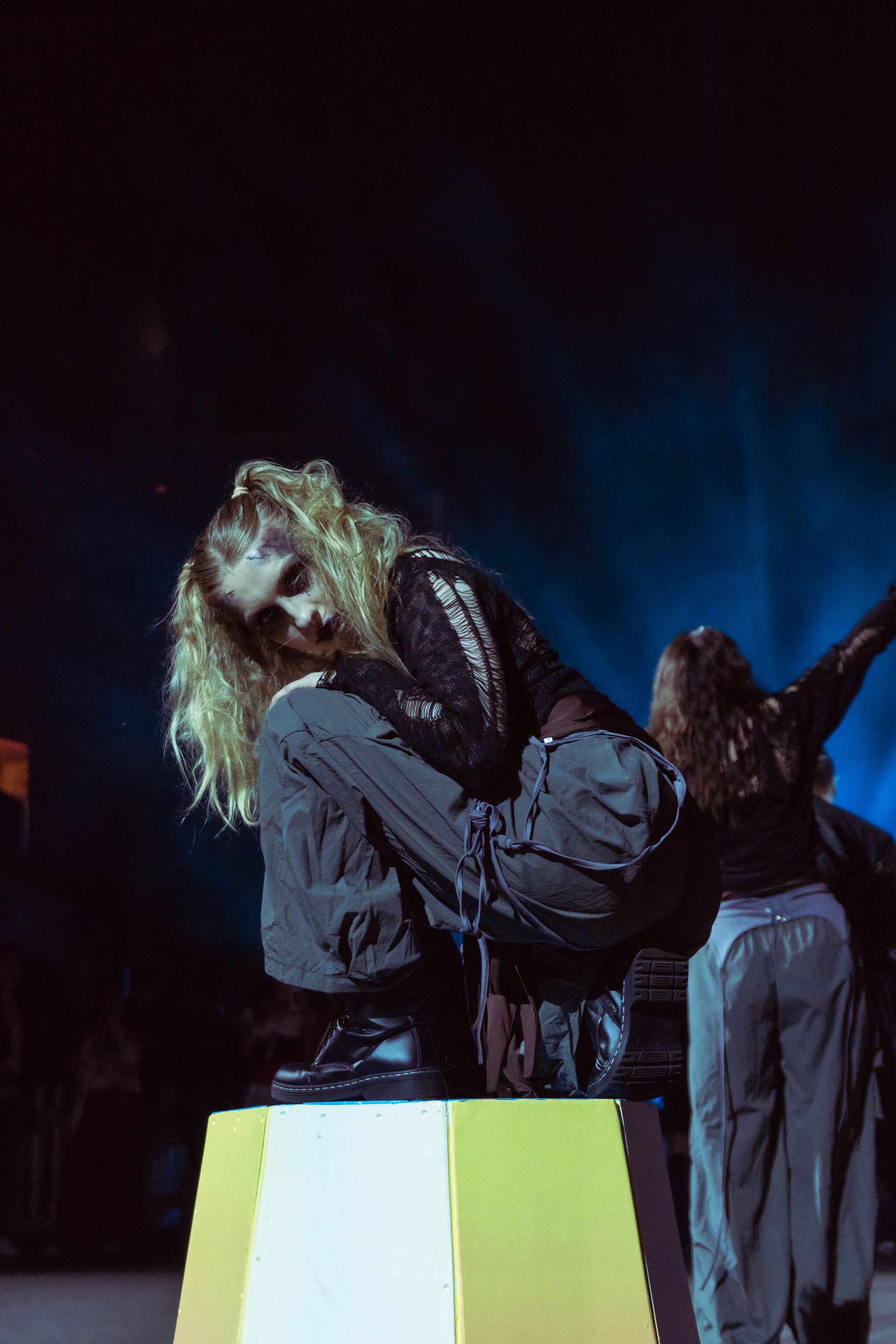 Performer with green hair crouching on a yellow box under dramatic blue stage lights at a live concert.