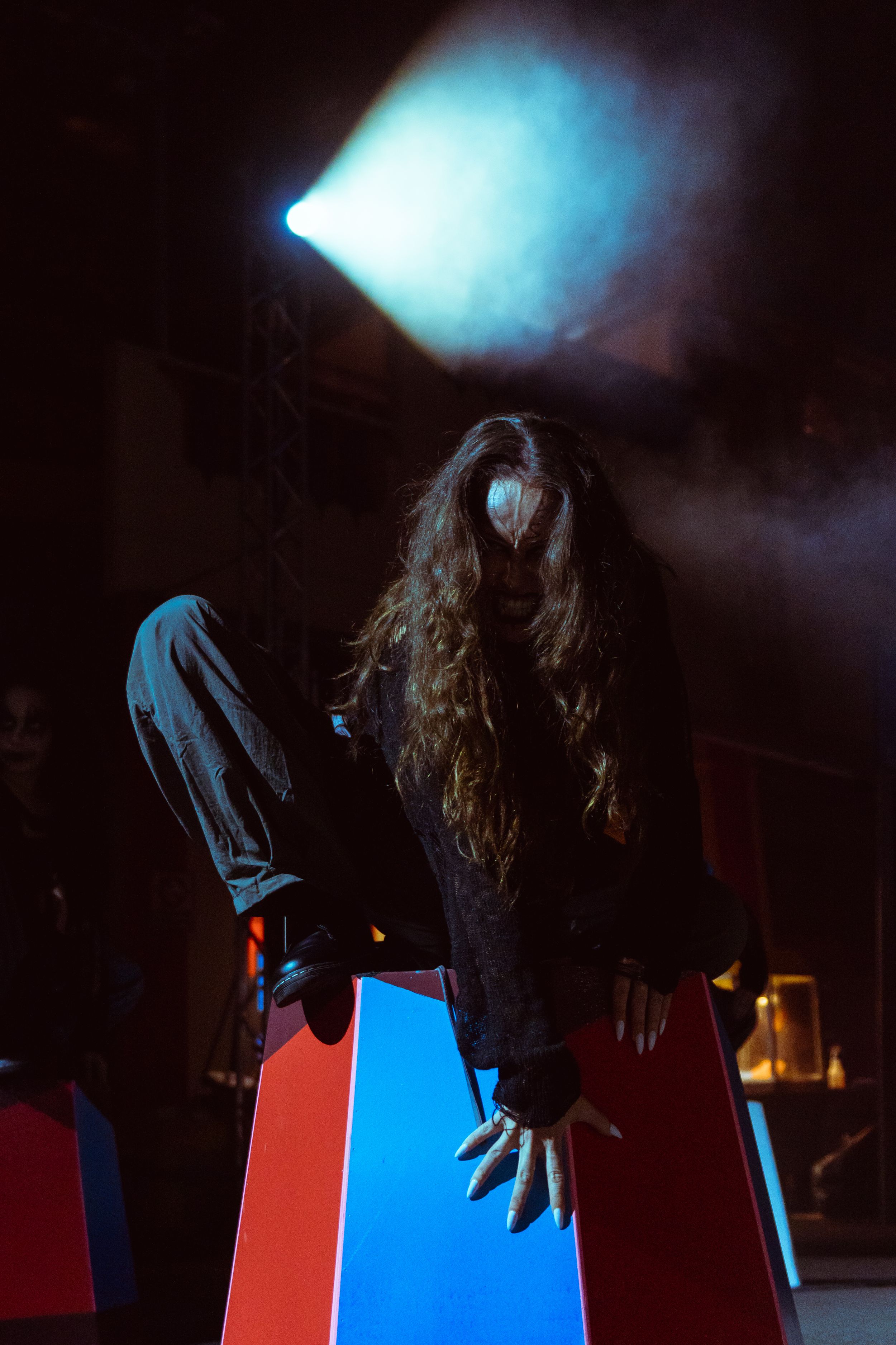 Creepy long-haired figure in dark costume crouching on colorful blocks under dramatic stage lighting.