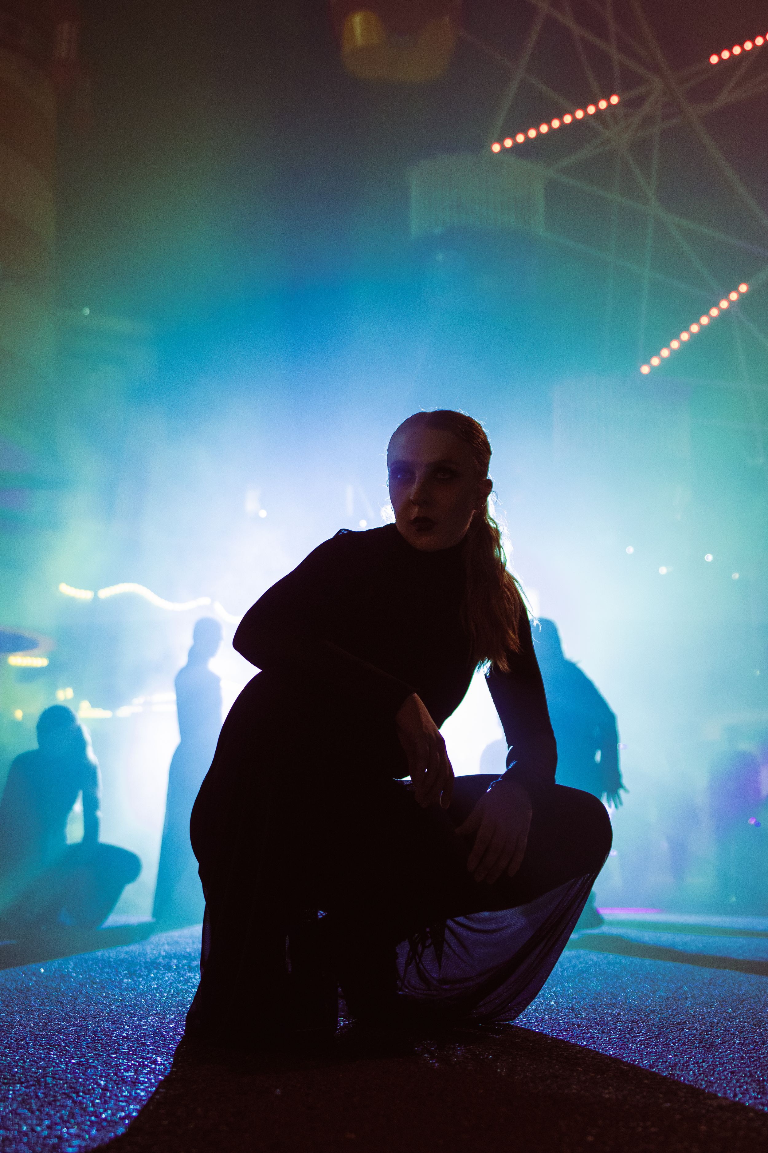 Person crouching on a dance floor surrounded by blue and green fog lights at a nightclub event.