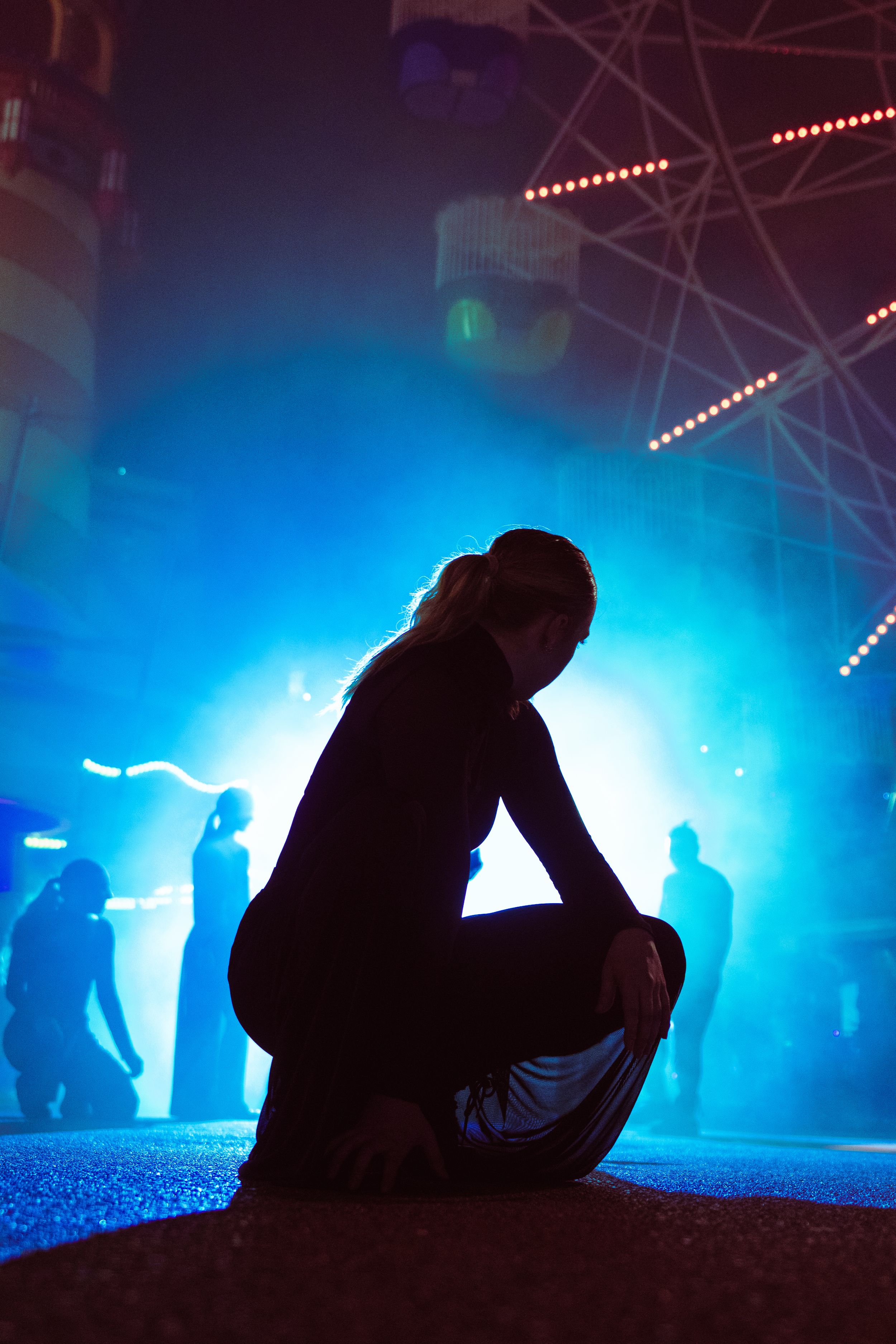Silhouette of a person crouching on a dance floor with blue stage lighting and crowd in background.