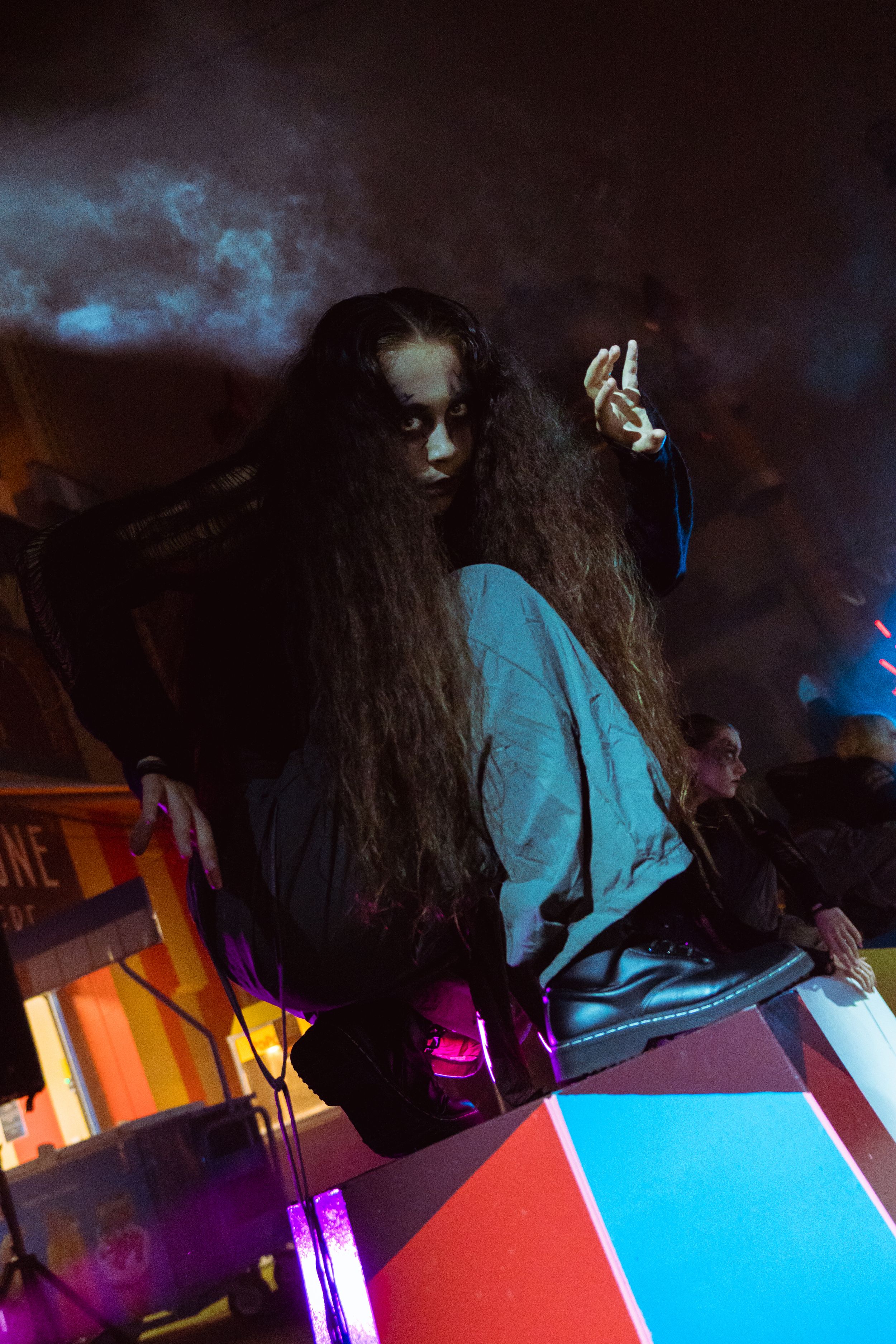 Woman with long curly hair in blue jacket poses dramatically at night event with colorful lights and smoke.