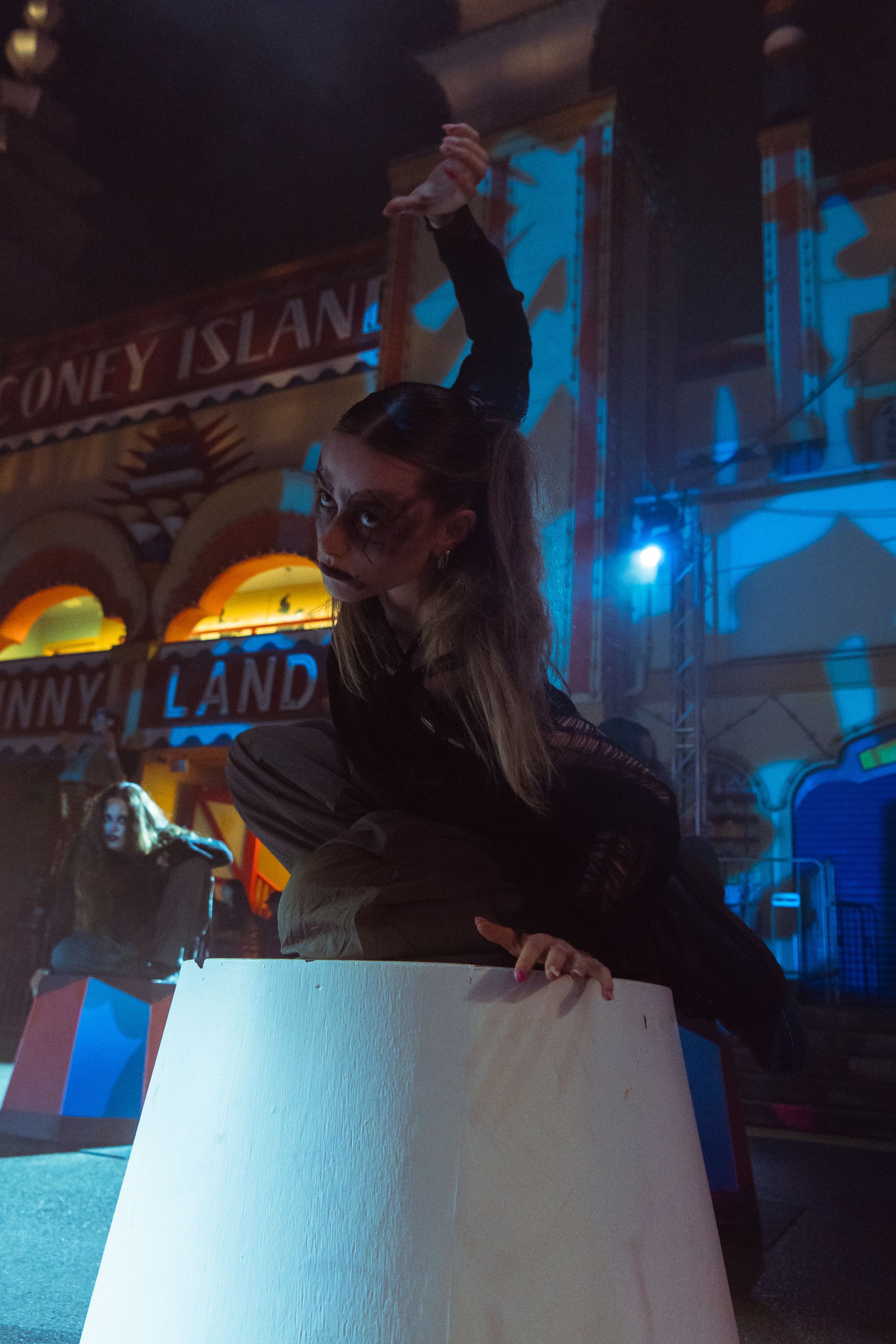 Woman in devil costume poses dramatically at Coney Island amusement park at night with blue lighting.