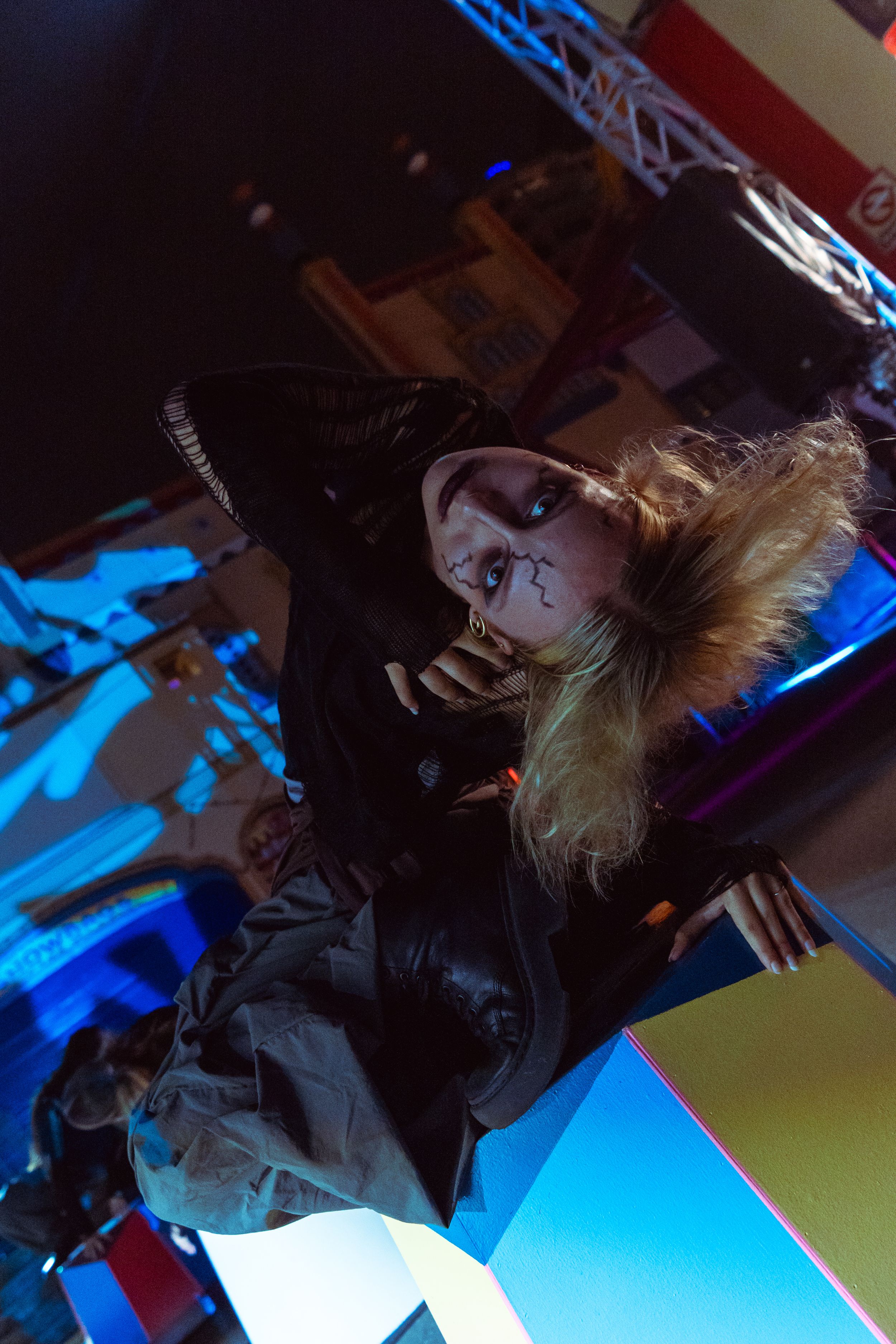 Woman with blonde hair and spider face paint at a nightclub event under colorful stage lights.