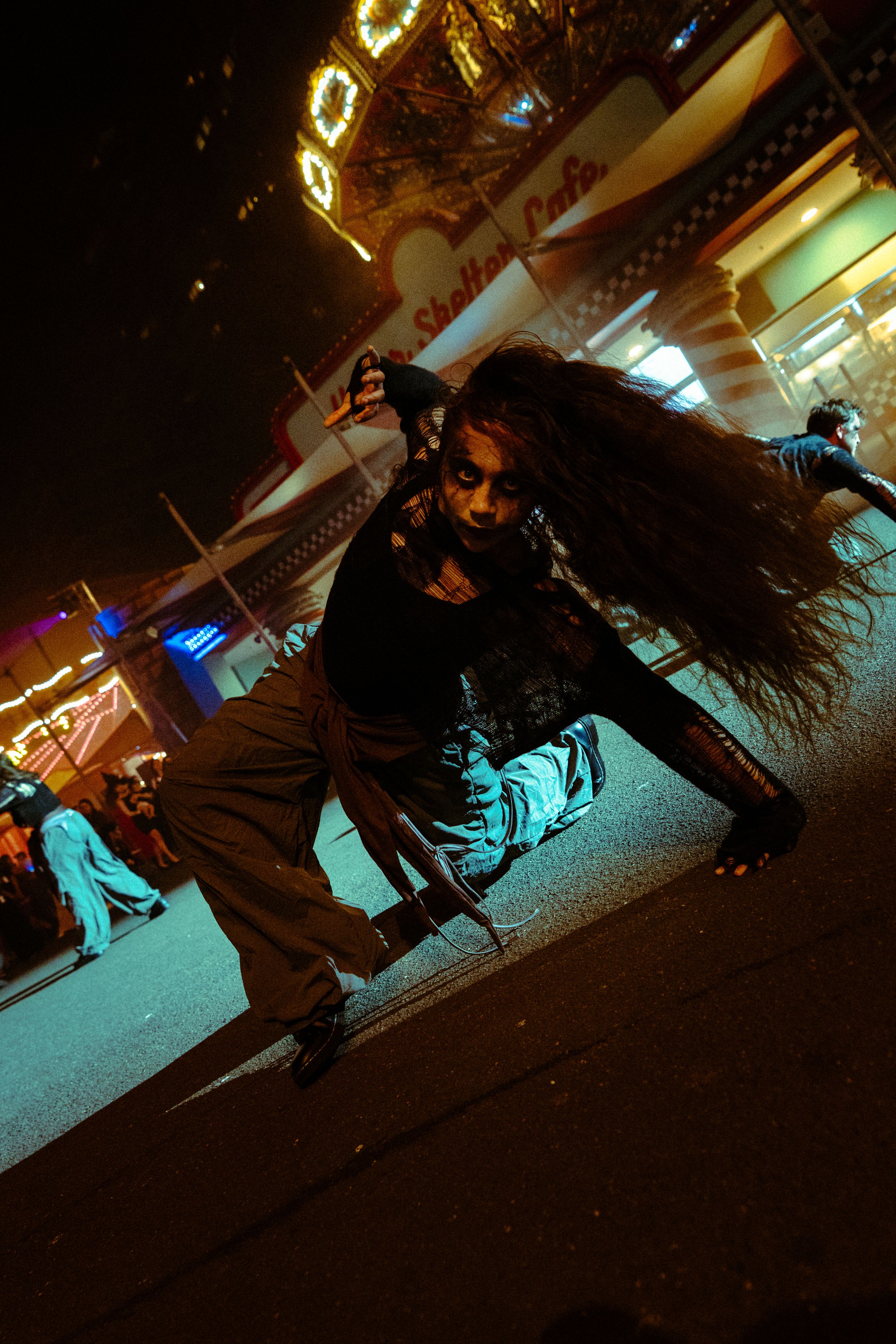 A zombie-costumed performer crawls on a dark street at a Halloween horror night event with neon lights in the background.