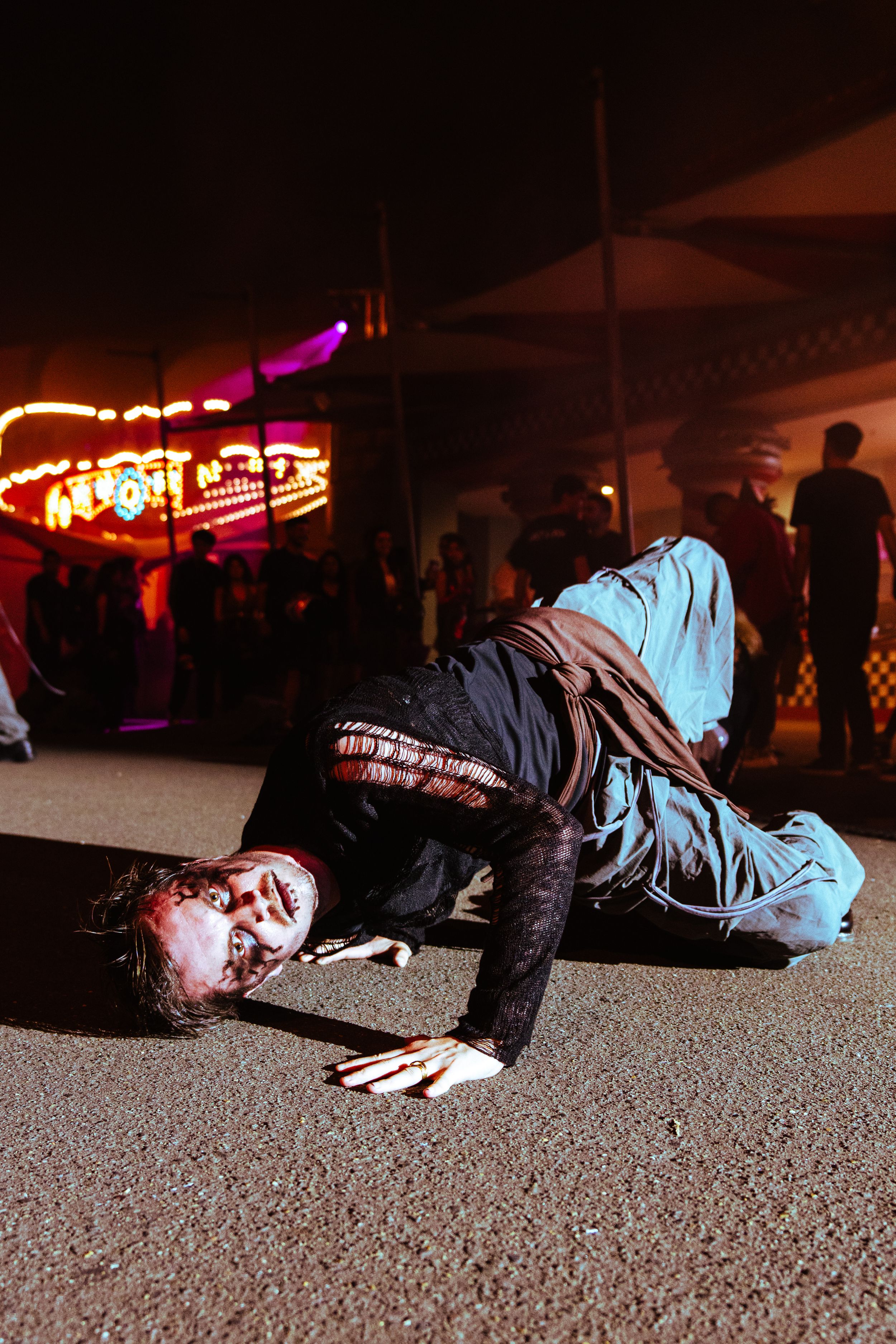 A male performer in zombie makeup crawls on the ground at a nighttime Halloween festival event.