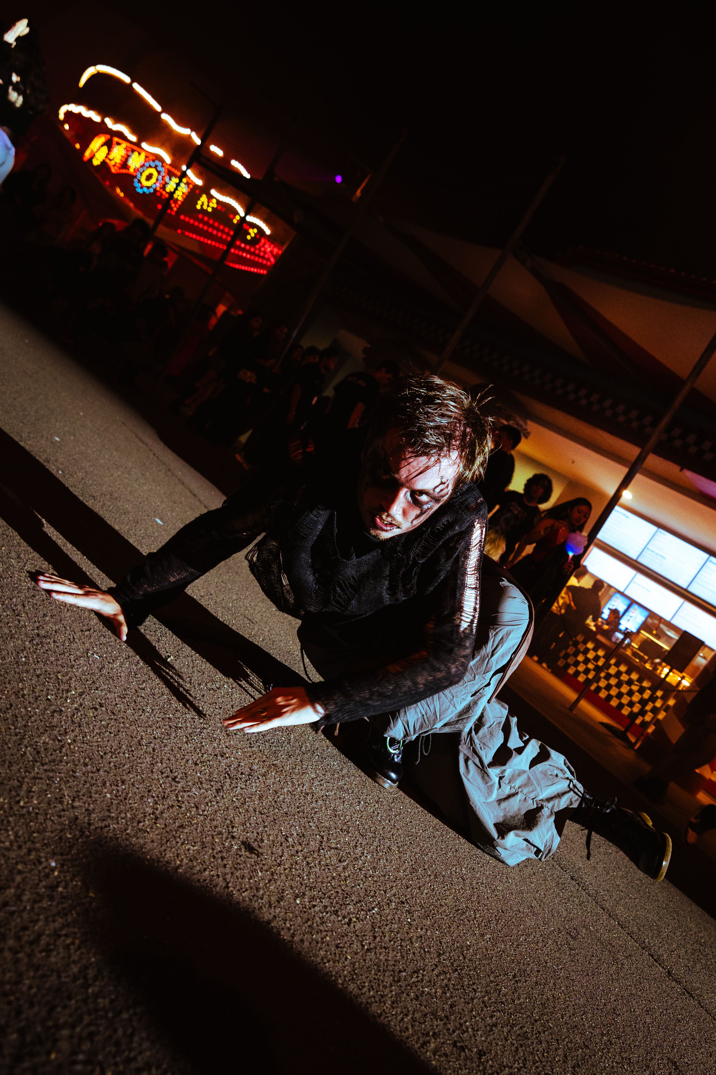 A zombie costume performer crouches dramatically on pavement at an outdoor Halloween night event.