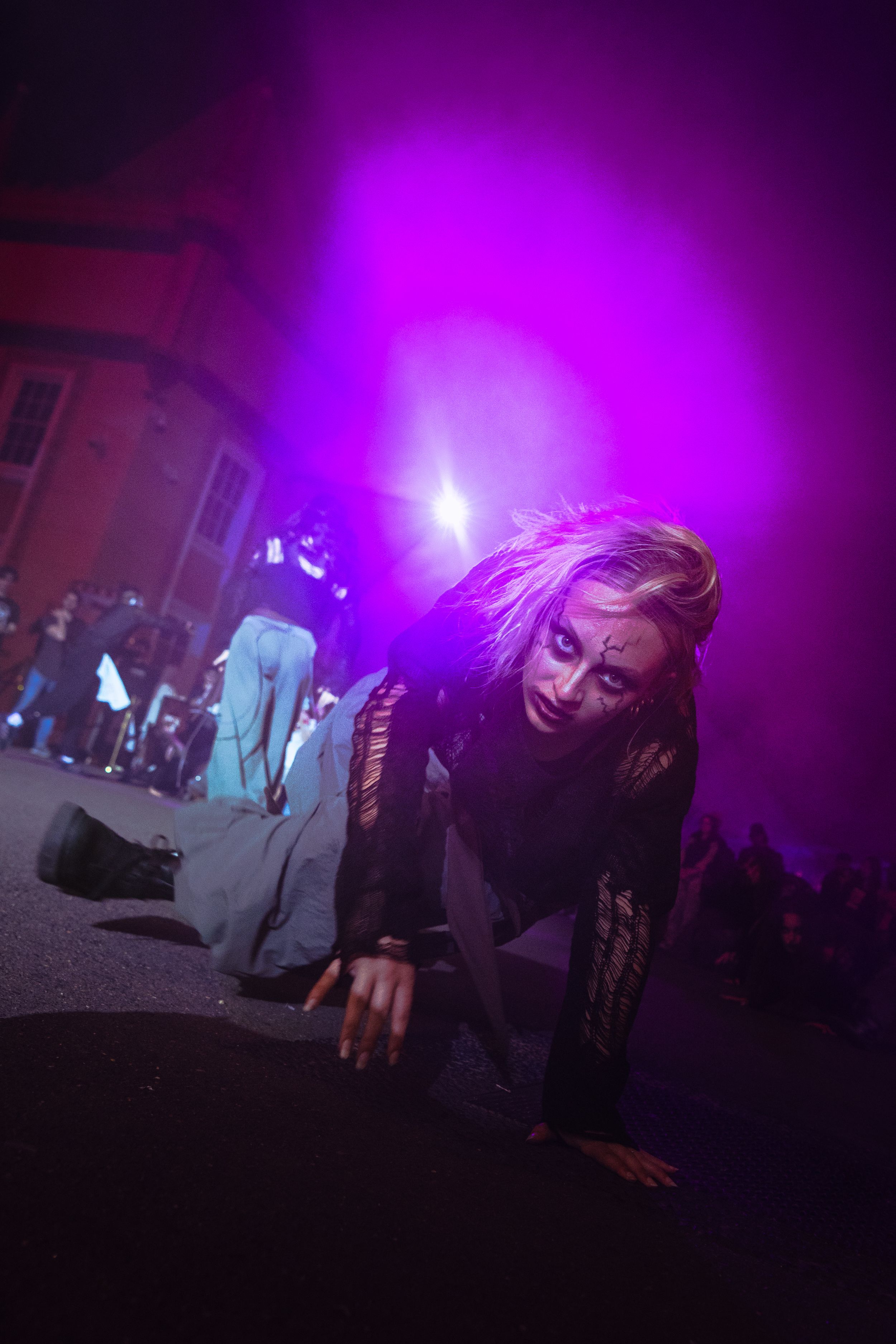A woman in zombie makeup crawls on the ground under purple lights at a Halloween street performance.