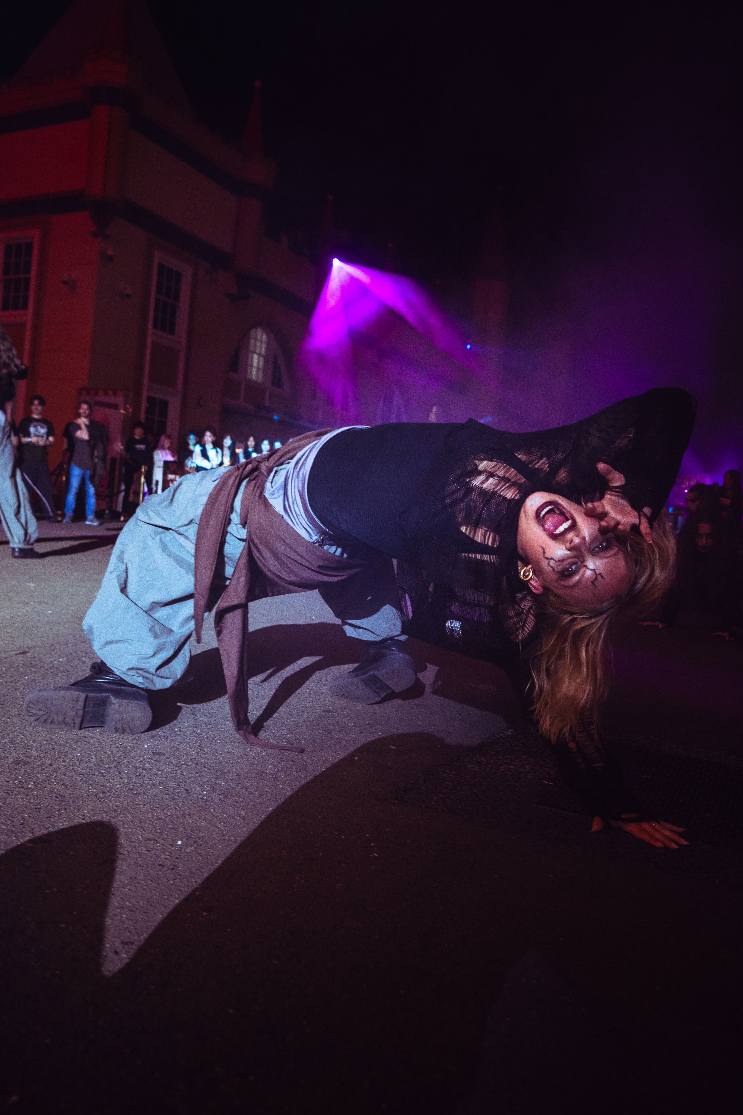 A Halloween performer in zombie makeup and witch hat poses low to the ground under purple stage lights.