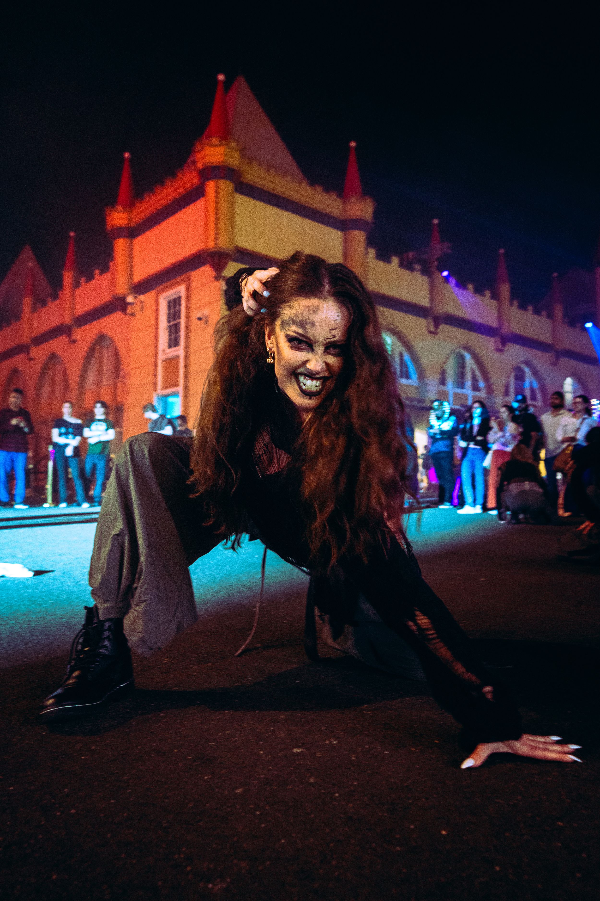 A woman with zombie face paint and long hair crouches menacingly at a Halloween night street event.