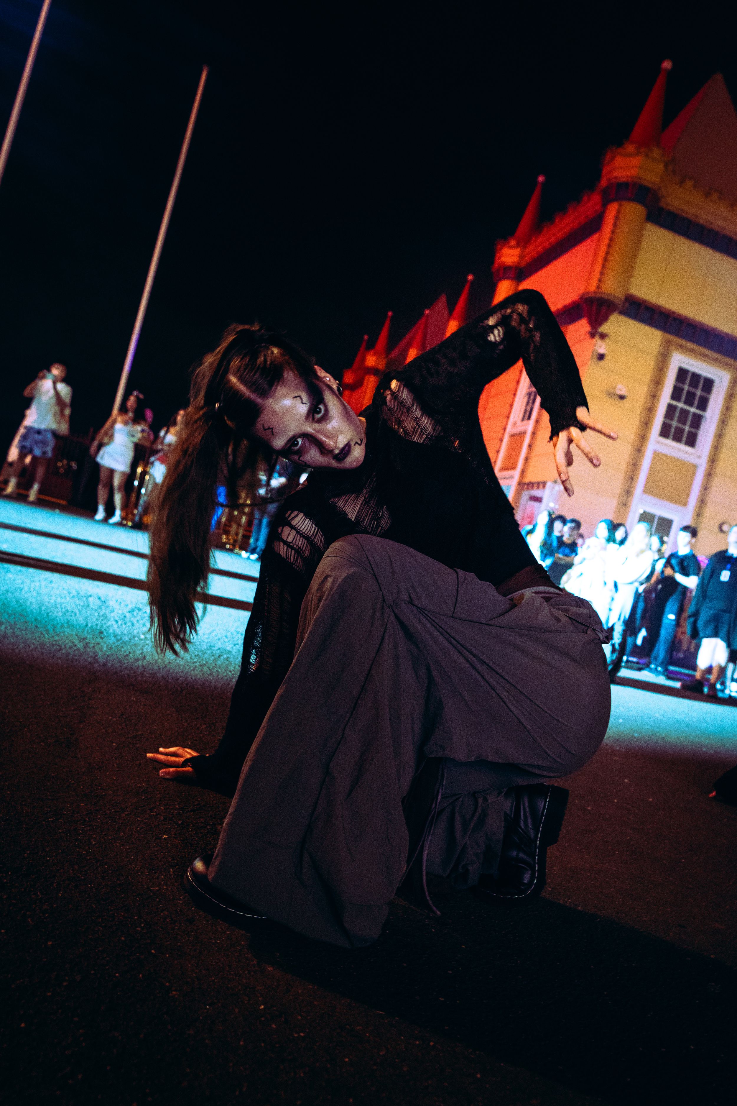 A female performer in dark zombie makeup sits on the ground gesturing dramatically at a night festival.