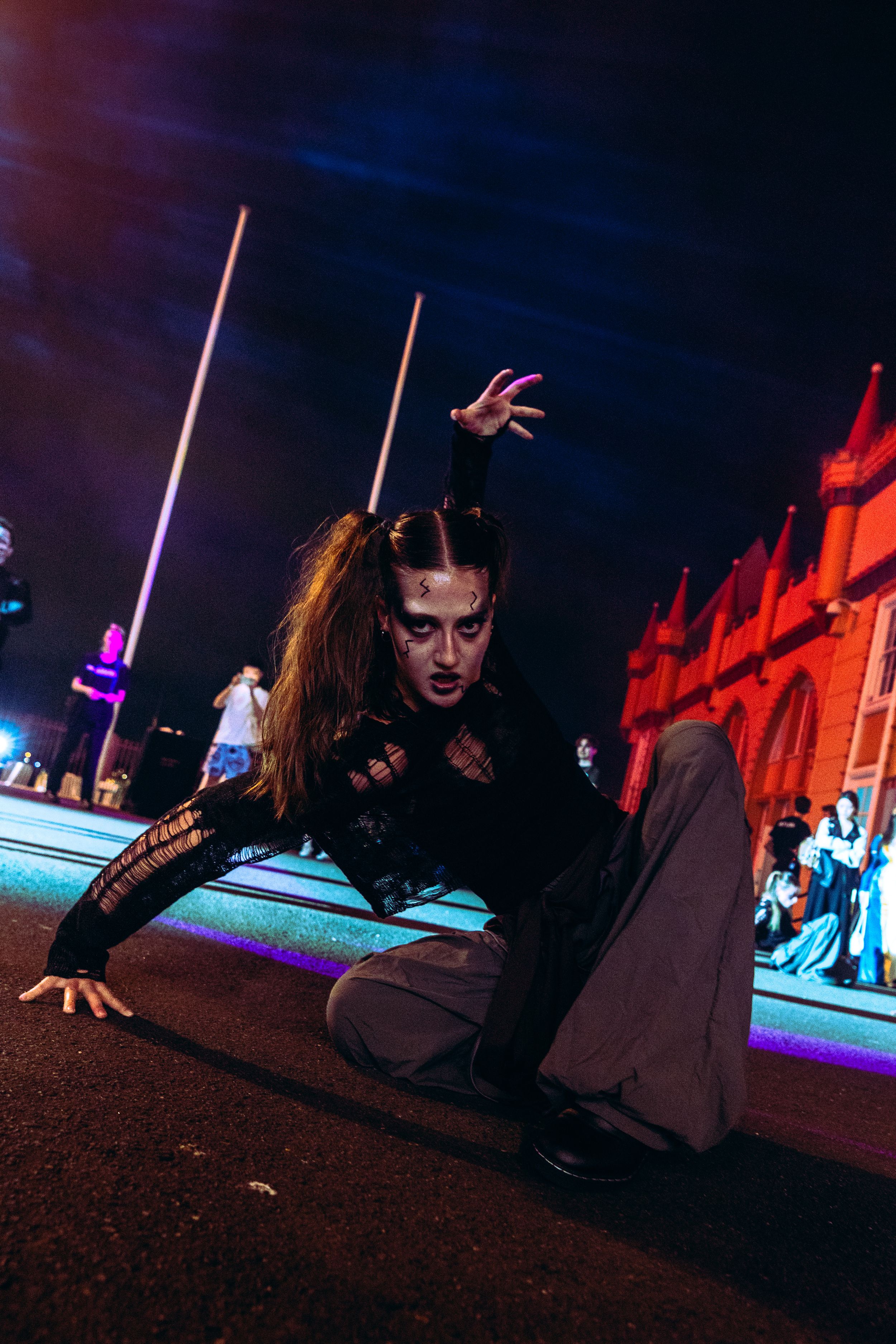 A zombie-costumed performer with devil horns crouches low on a lit street at a Halloween night event.