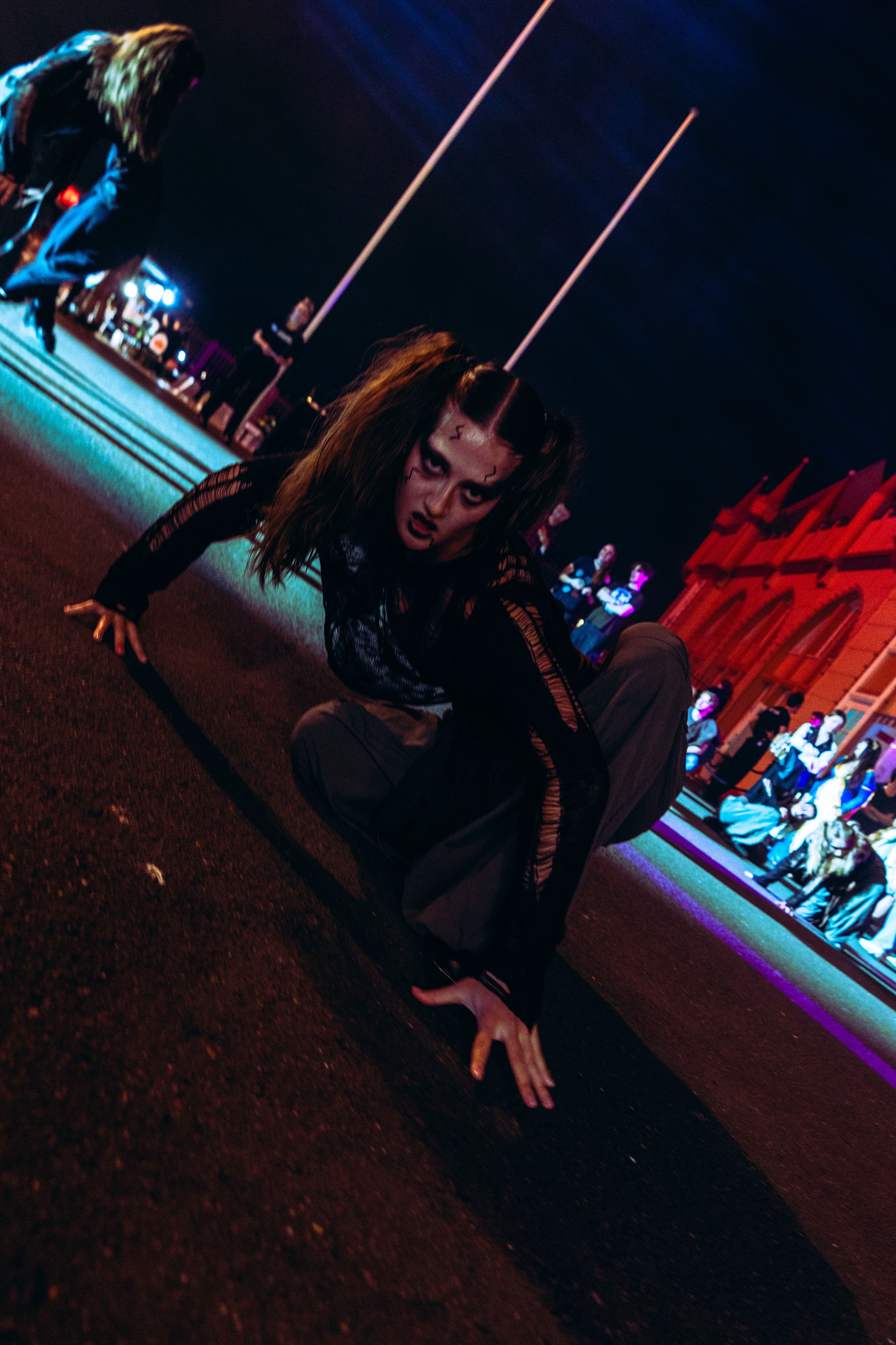 A Halloween performer in dark costume and zombie makeup crouches on pavement at a nighttime festival.