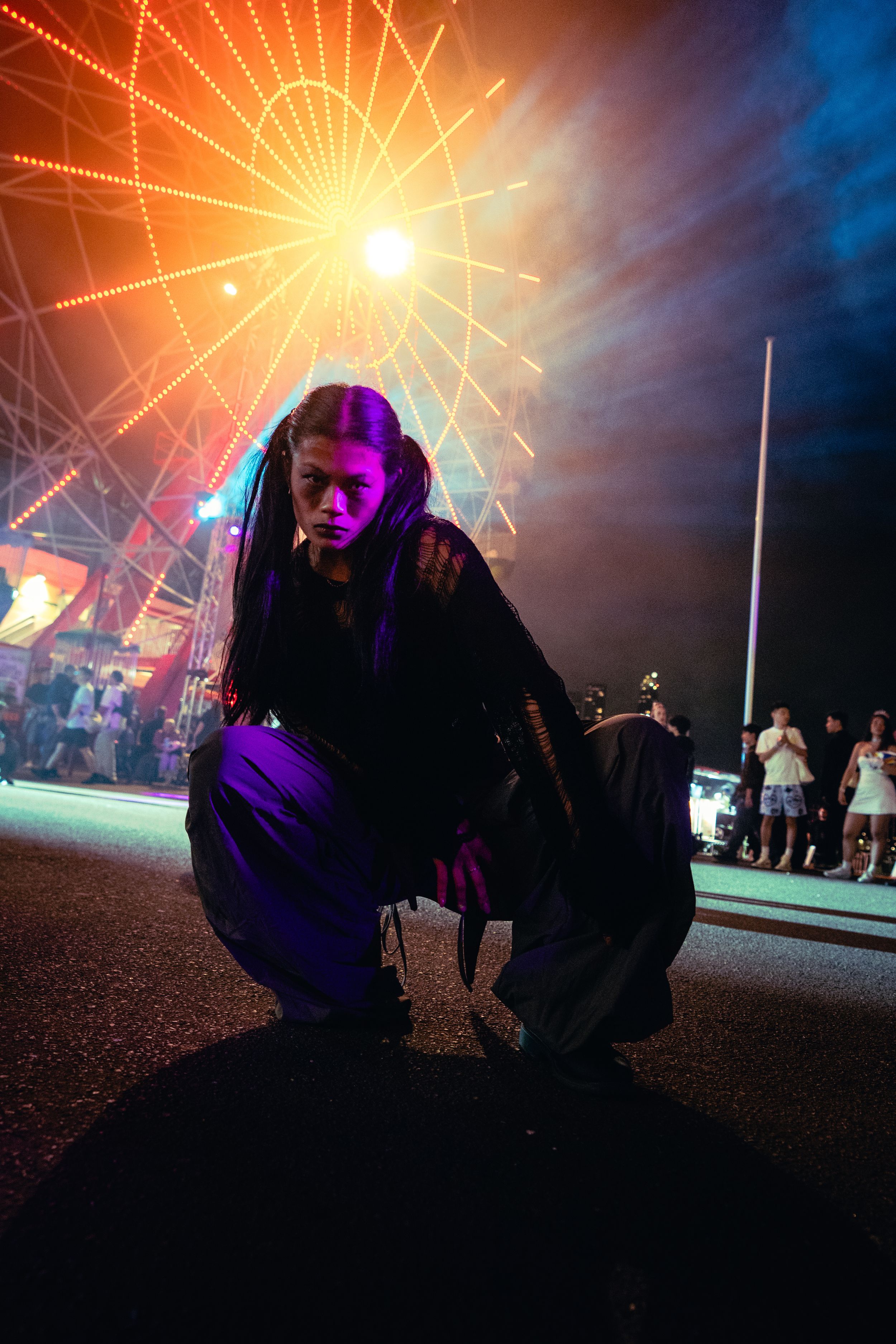 A Halloween performer in dark costume crouches under colorful lights with a Ferris wheel in background.