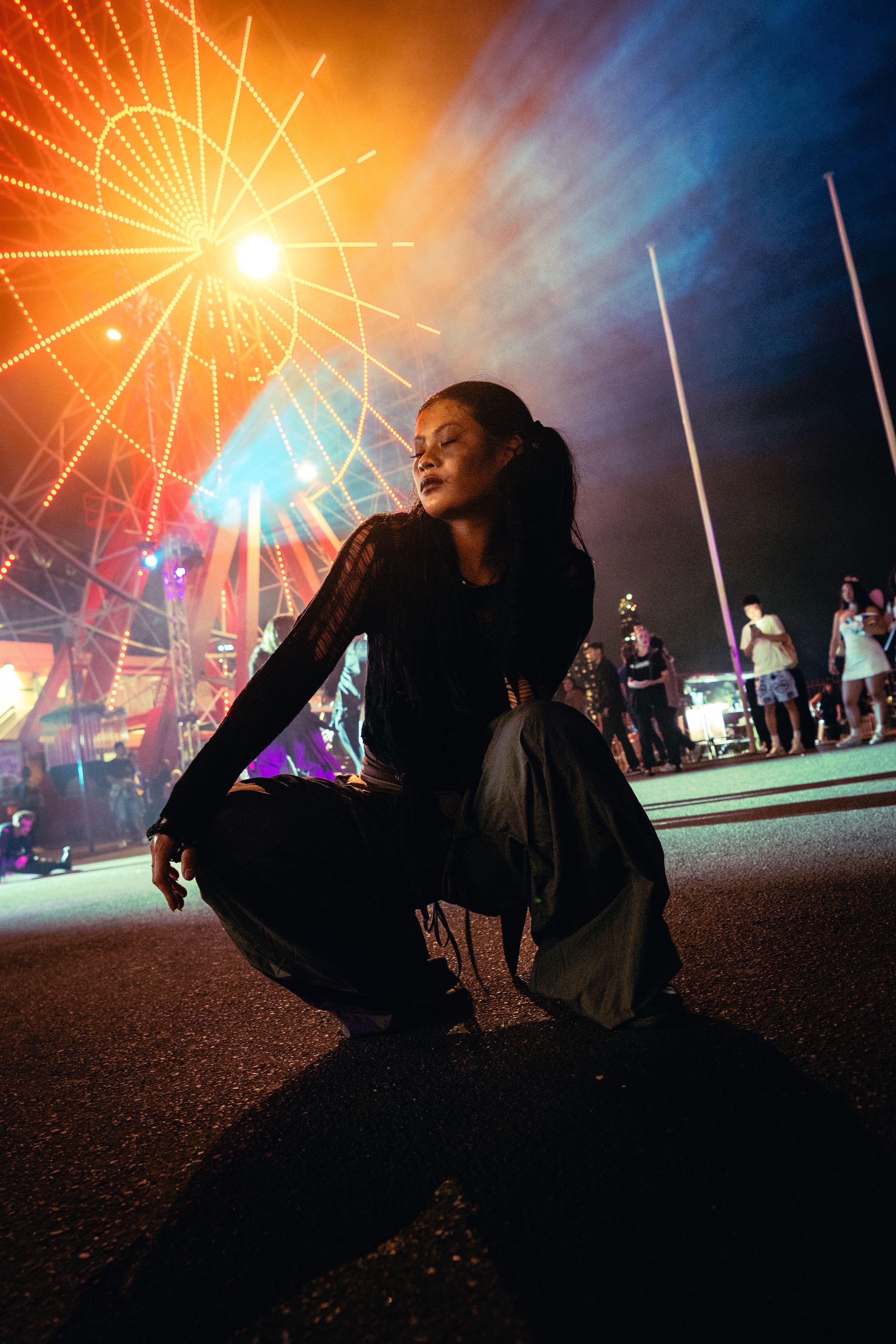 A woman in black crouches gracefully on a street with a glowing Ferris wheel at a night carnival.