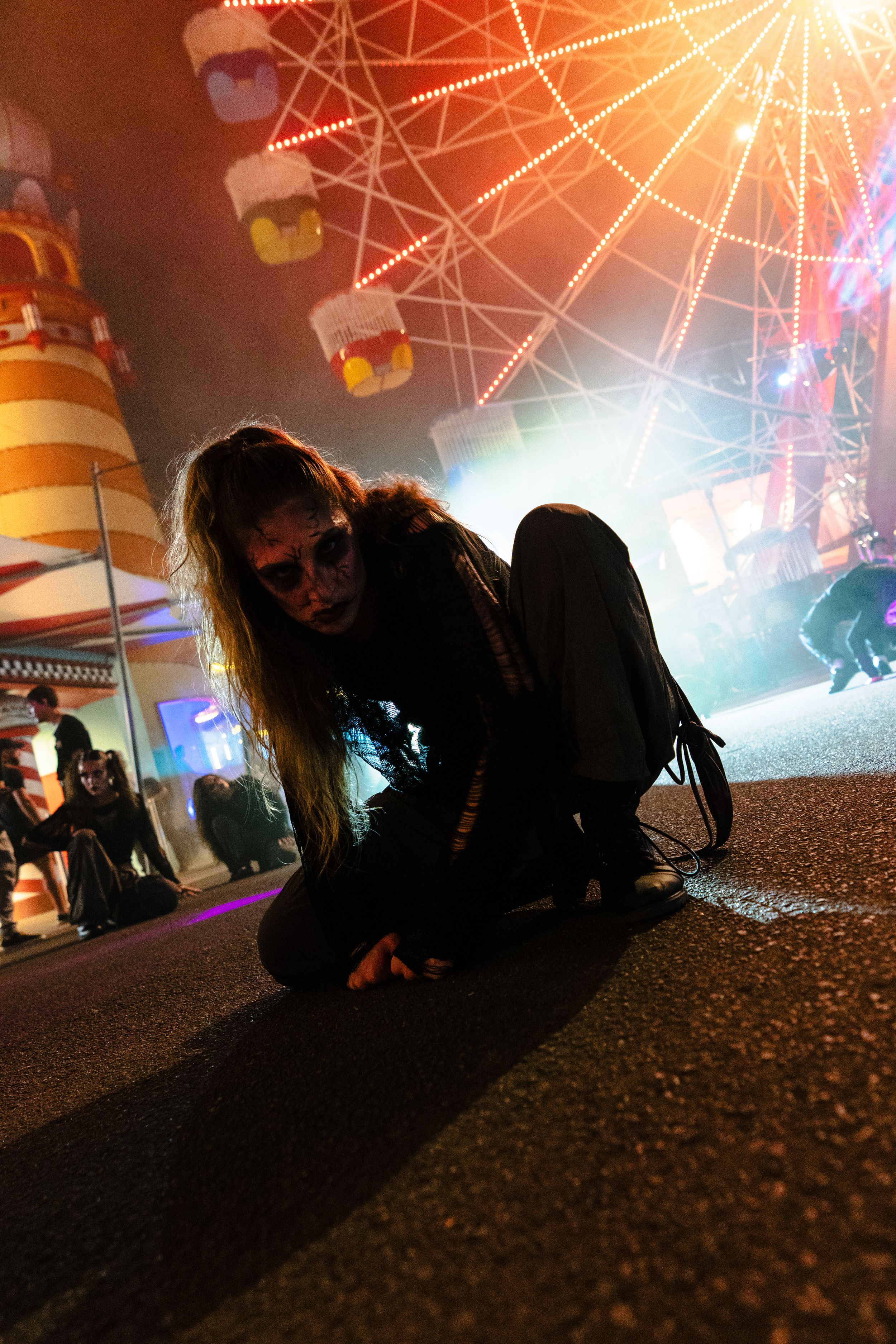 Woman in zombie makeup crawls on pavement at night carnival with ferris wheel lights glowing behind her.