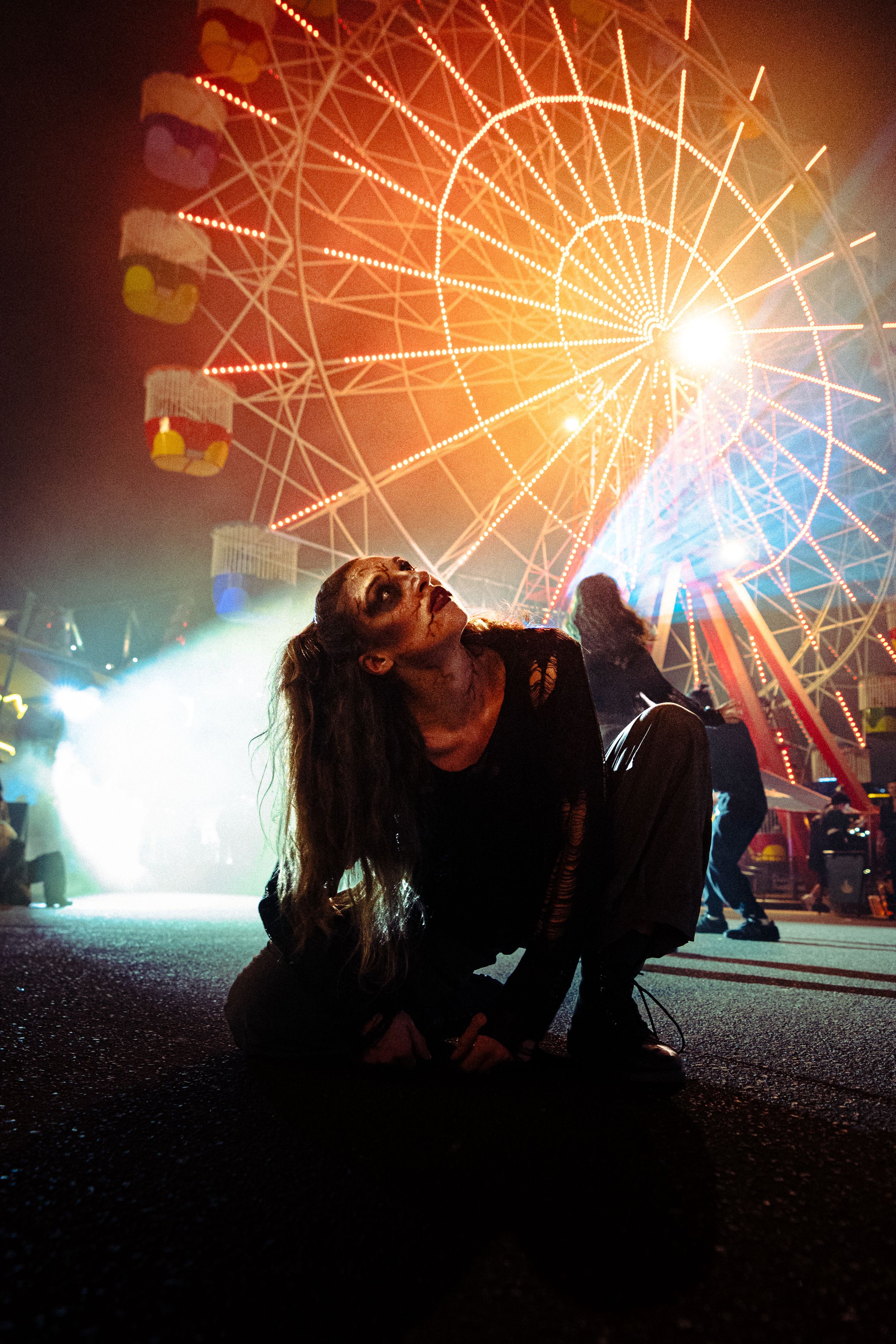 Woman with zombie face paint kneels on pavement before illuminated ferris wheel at Halloween carnival.