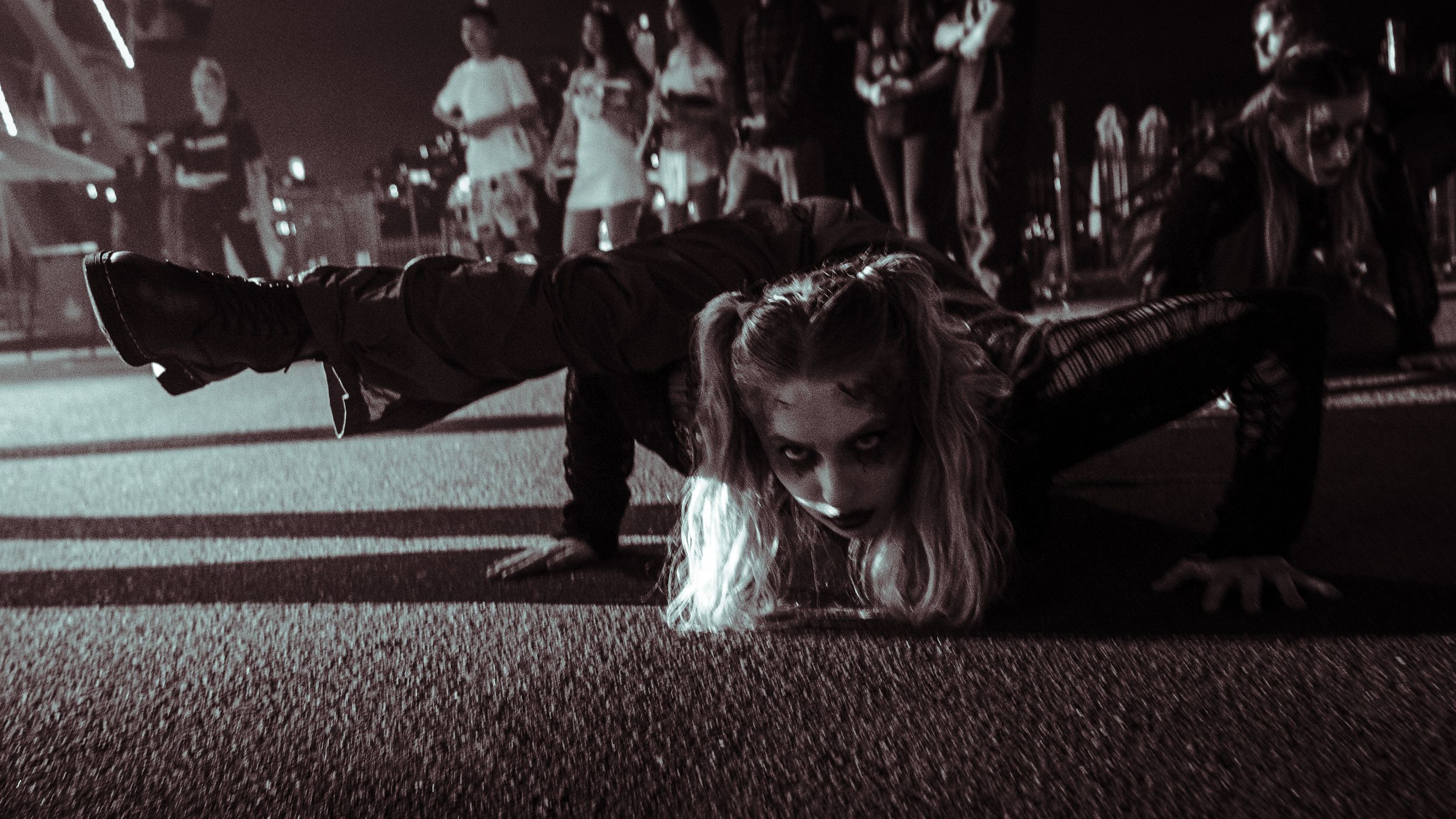 Person in zombie costume crawling on ground at dark outdoor Halloween event with crowd watching.