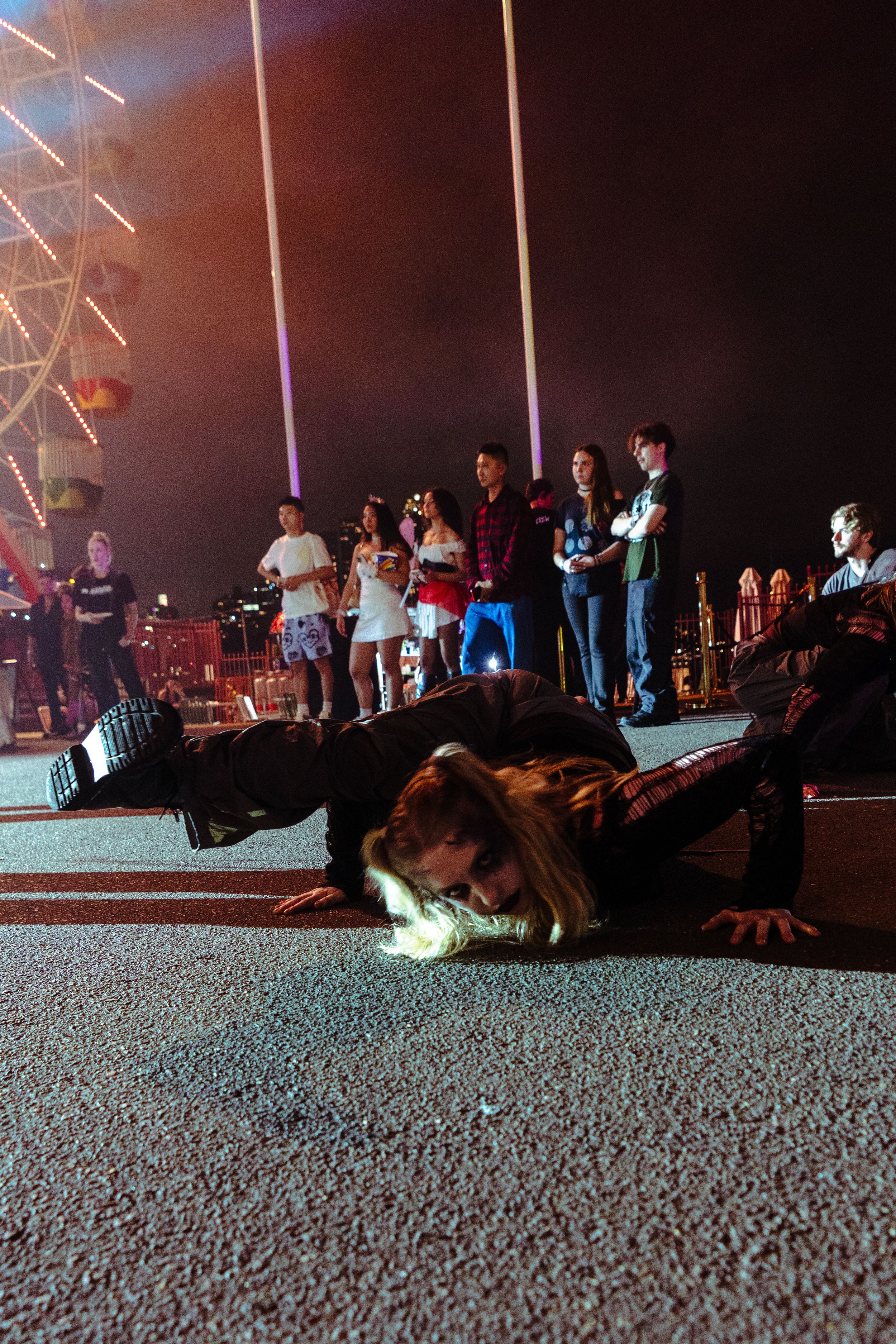 Zombie performer does push-ups on pavement at night carnival while crowd watches near ferris wheel.