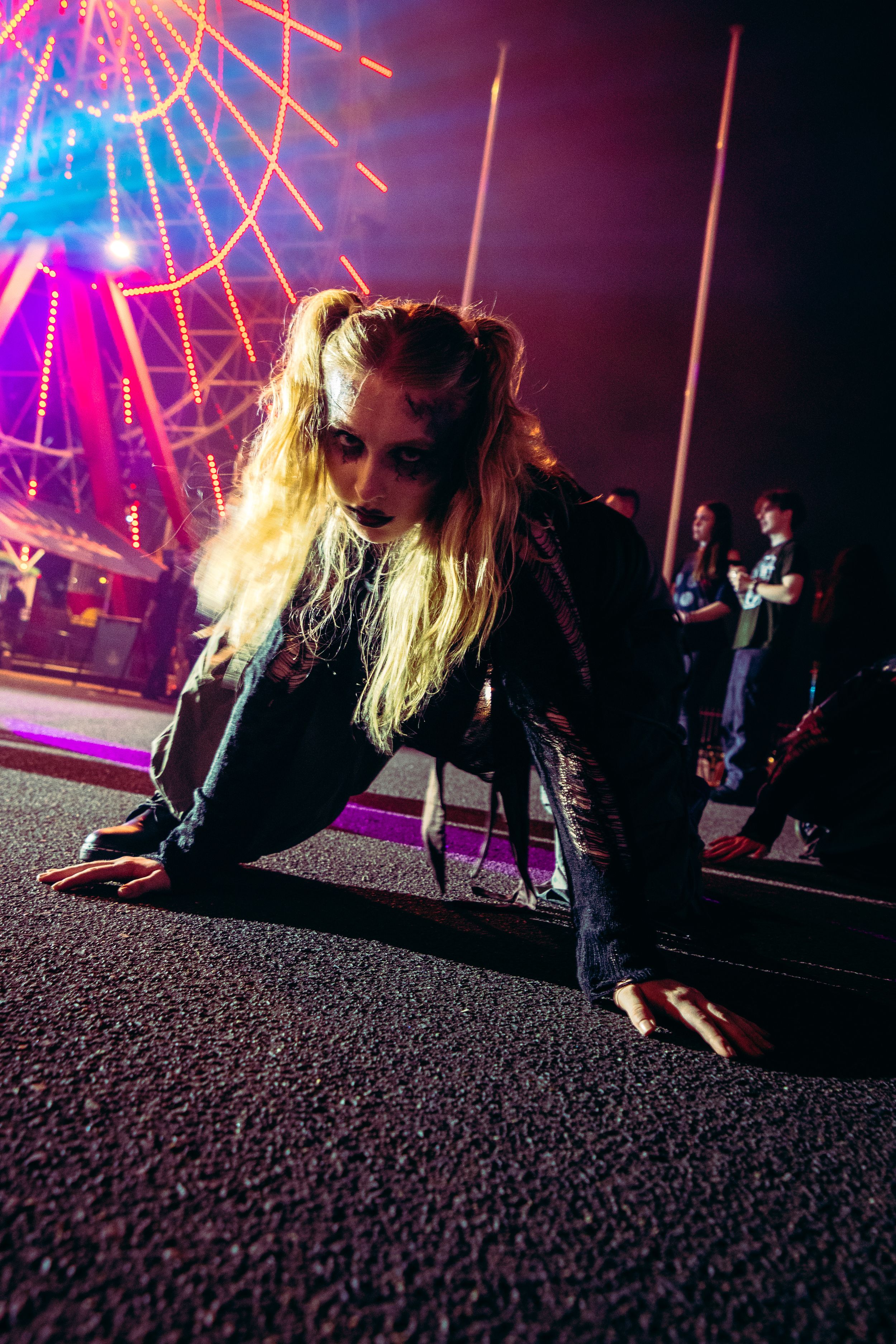 Blonde woman in zombie makeup crawls on asphalt at colorful Halloween carnival with ferris wheel.