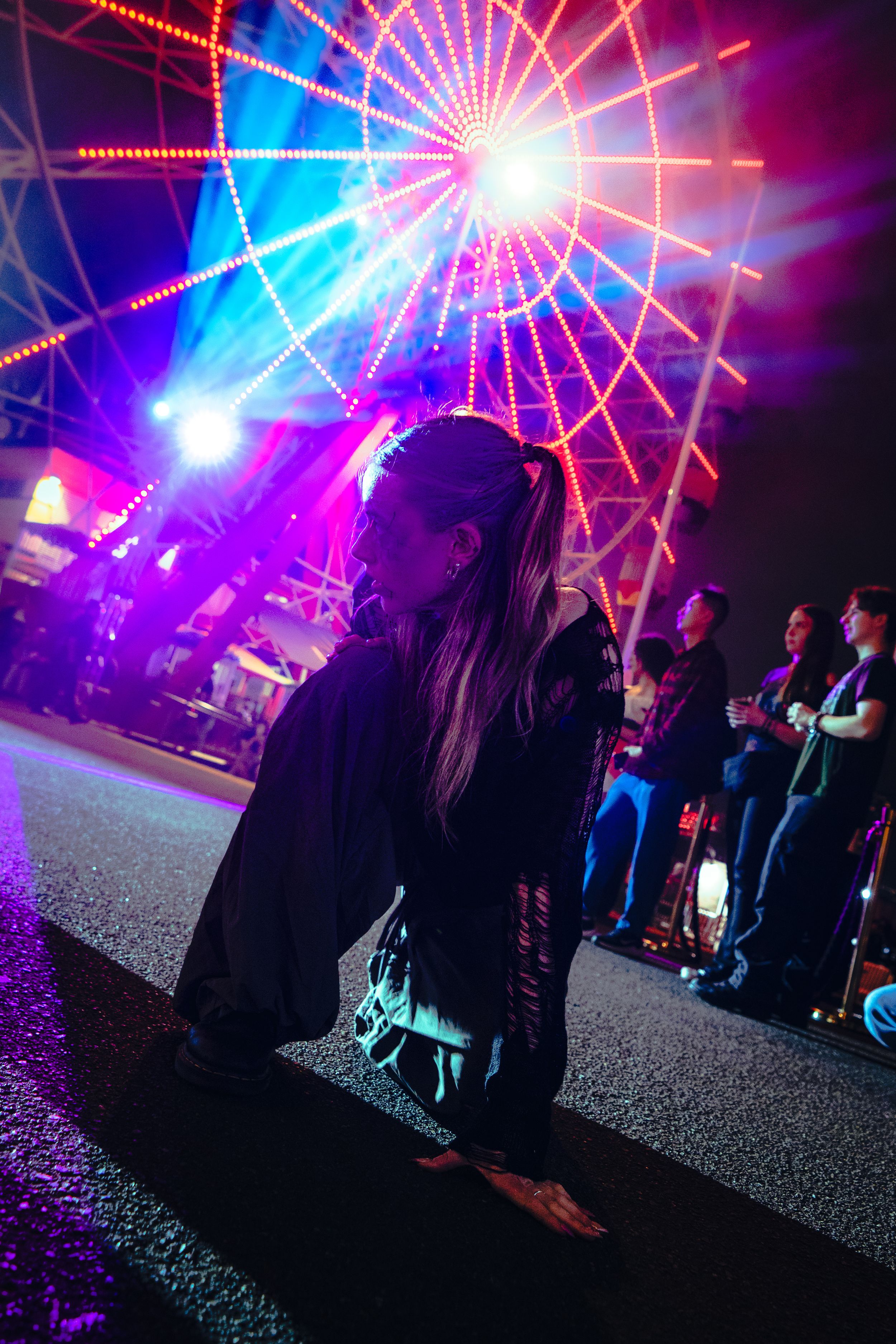 Woman in dark costume kneels on street at vibrant Halloween carnival with lit ferris wheel.